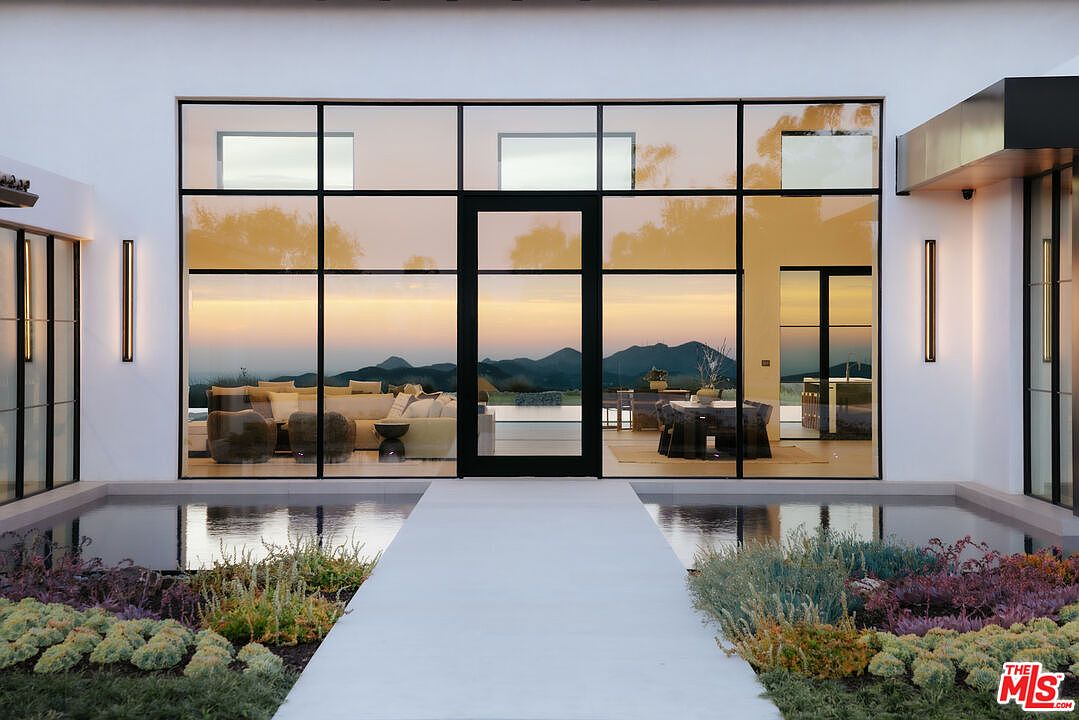 This striking modern entryway features a dramatic glass-walled facade that offers a seamless view into the home's interior living and dining spaces. A minimalist concrete walkway extends across a reflective water feature, leading directly to the central glass door, while lush, low-profile landscaping frames the path. The composition emphasizes clean lines, architectural symmetry, and a sophisticated connection between the indoor and outdoor environments.