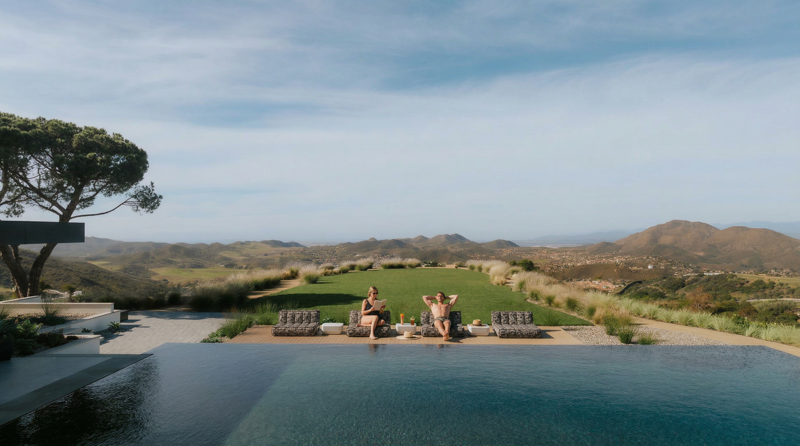 This stunning infinity-edge pool overlooks a vast, rolling landscape of hills and valleys, creating a seamless visual connection between the water and the horizon. A manicured lawn sits between the pool deck and the natural terrain, accented by ornamental grasses and a row of modern lounge chairs. The perspective is captured from a slightly elevated vantage point, emphasizing the luxurious, serene atmosphere of this high-end outdoor living space.