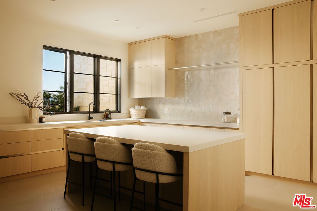 This modern, minimalist kitchen features light wood cabinetry, a large central island with seating, and a sleek, neutral color palette. The space is illuminated by natural light streaming through a large window, highlighting the clean lines and high-end finishes. The overall aesthetic is bright, airy, and sophisticated, perfect for contemporary living.