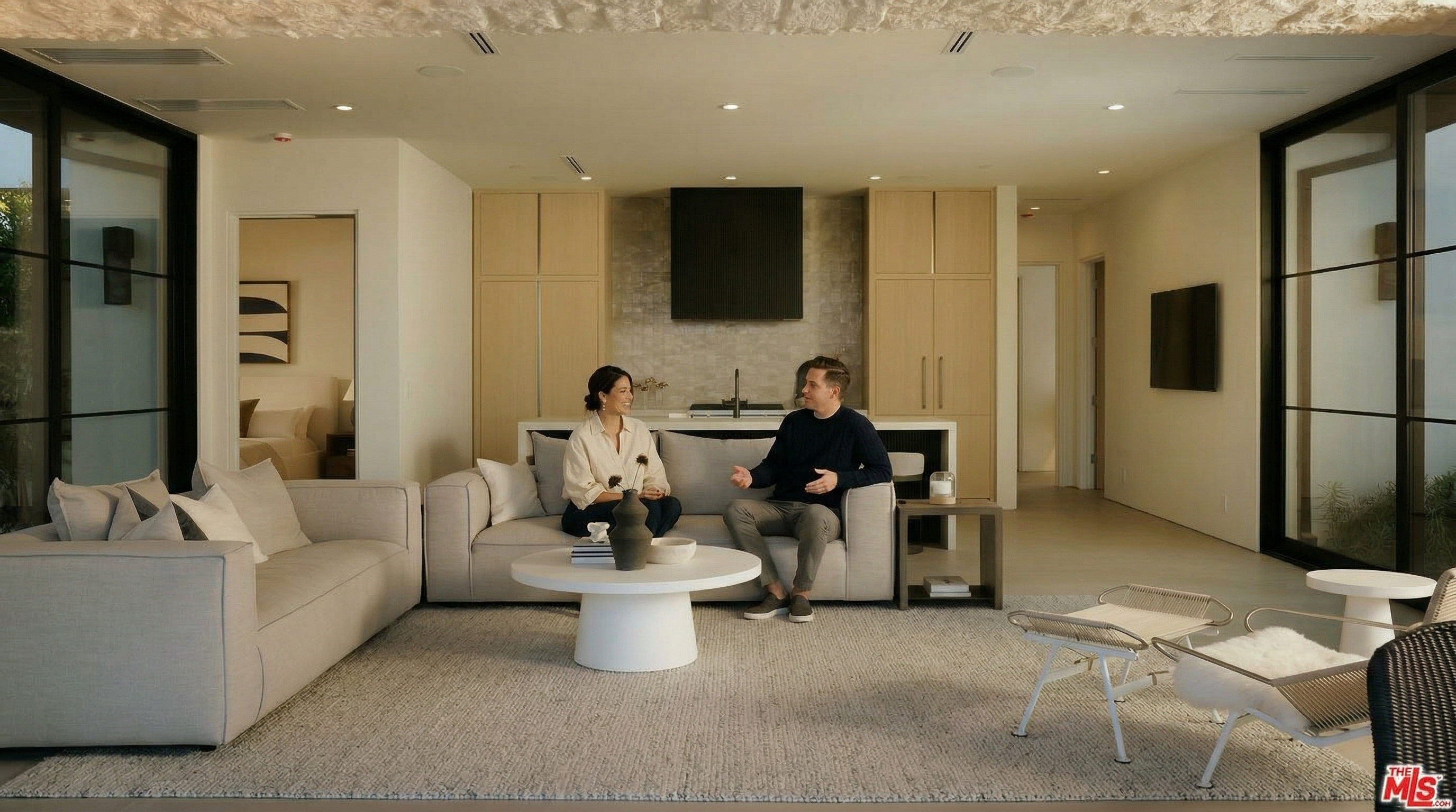 This sophisticated open-concept living area features a neutral color palette, plush contemporary sofas, and a minimalist round coffee table centered on a textured area rug. The space is anchored by a sleek kitchen island in the background, framed by light wood cabinetry and a textured stone wall, creating a seamless blend of comfort and modern luxury. The perspective is a wide, eye-level shot that emphasizes the airy, high-end design and the cohesive flow between the living and kitchen zones.