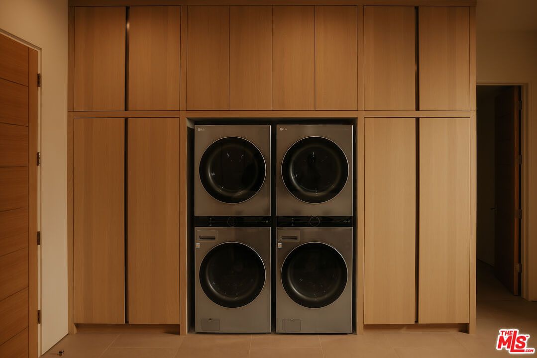 This modern laundry room features a sleek, minimalist design with light wood cabinetry that seamlessly integrates a stacked LG washer and dryer unit. The clean lines and neutral color palette create a sophisticated and organized atmosphere, emphasizing high-end functionality. The perspective is a straight-on, eye-level shot that highlights the symmetry and custom built-in storage solutions.