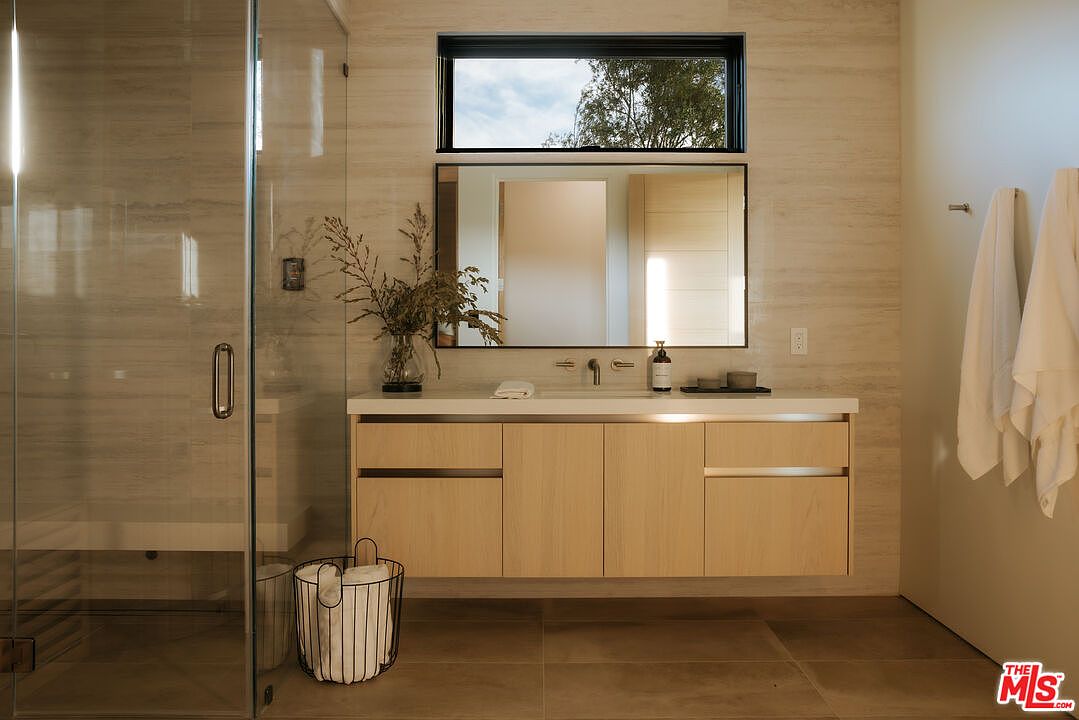 This modern primary bathroom features a floating light-wood vanity with a sleek countertop and a large, rectangular mirror centered beneath a horizontal window. To the left, a glass-enclosed walk-in shower adds to the clean, minimalist aesthetic, while a wire basket with rolled towels sits on the floor. The space is finished with neutral, textured wall tiles and large-format floor tiles, creating a serene and contemporary atmosphere.