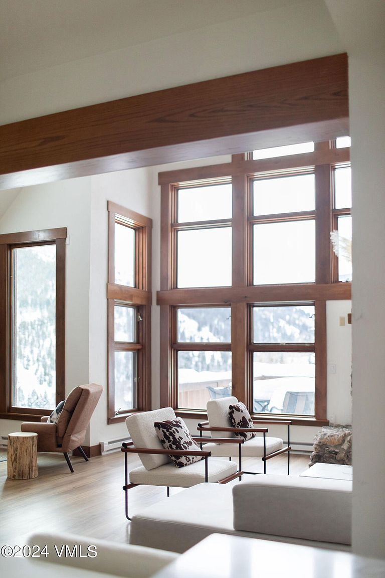 This living room features large windows with wooden frames that offer a view of a snowy landscape. The room is furnished with modern chairs and a sofa, creating a cozy and inviting atmosphere. A wooden beam adds an architectural detail to the space.