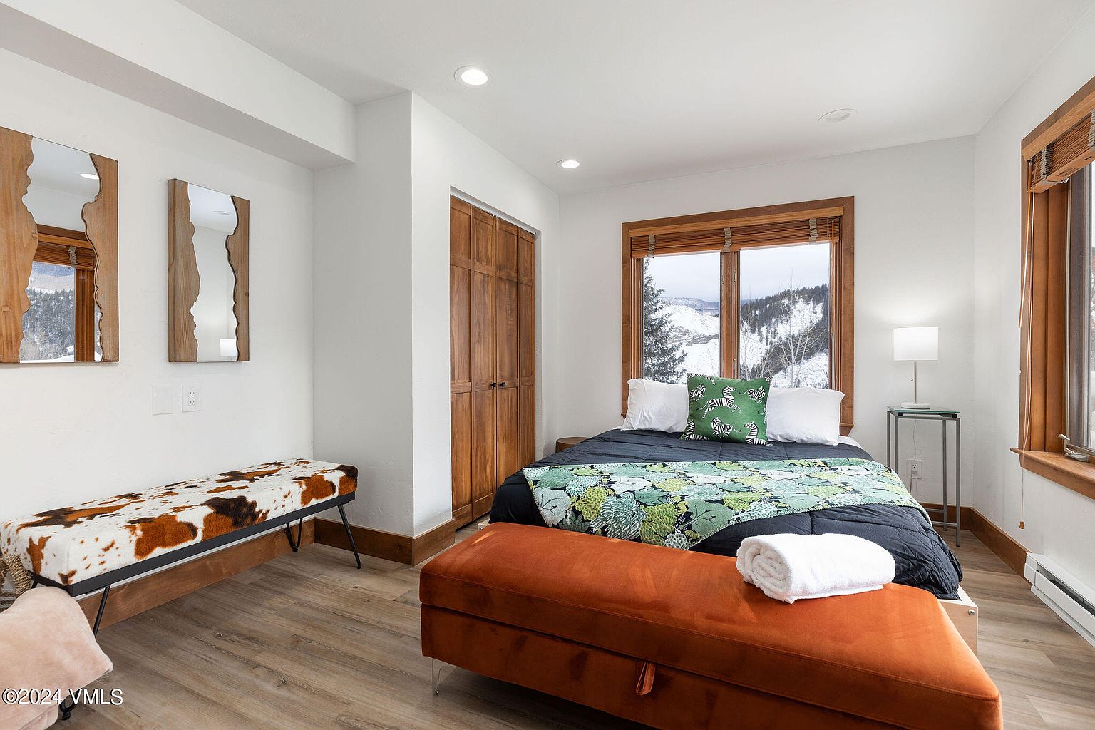 This is a cozy guest bedroom featuring a bed with a dark blue comforter and a green patterned throw, complemented by an orange velvet storage bench at the foot of the bed. The room has wooden-framed windows offering a view of a snowy landscape, and the decor includes unique wooden-framed mirrors on the wall. The flooring is light wood, and the overall impression is warm and inviting.