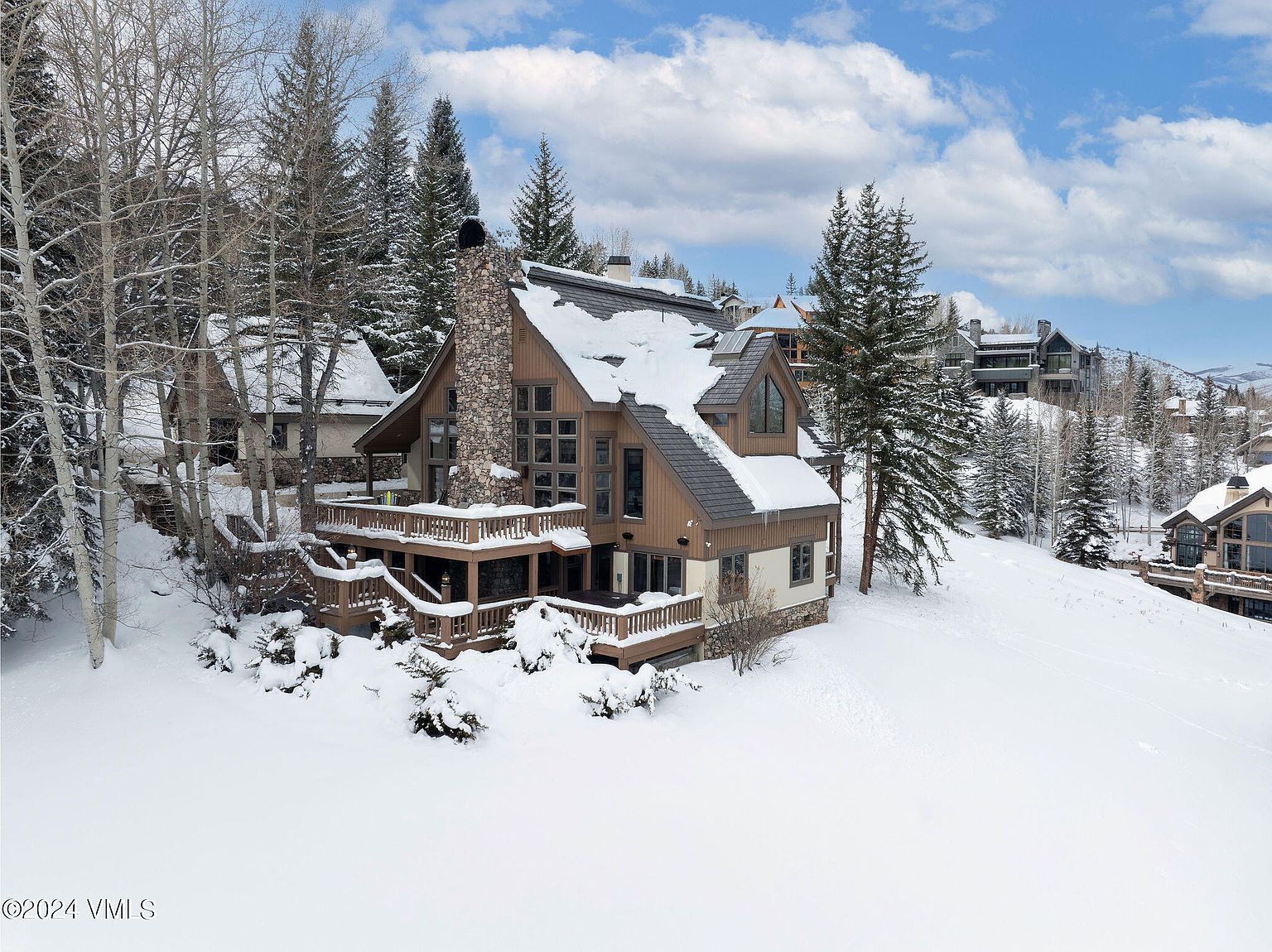 This aerial view showcases a luxurious mountain home nestled in a snowy landscape. The house features a stone chimney, multiple decks, and large windows, blending rustic charm with modern design. The surrounding trees and snow-covered terrain create a serene and picturesque setting, ideal for a winter retreat.