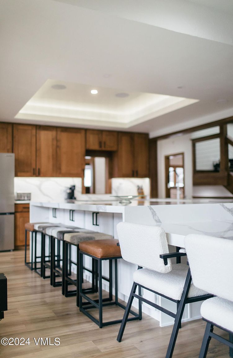 This interior shot showcases a modern kitchen with warm wood cabinetry and a large island featuring a white countertop with marble veining. The island is surrounded by a mix of bar stools with black frames and varying seat materials, including leather and textured white fabric. The kitchen is well-lit with recessed lighting and natural light, creating an inviting and stylish space.