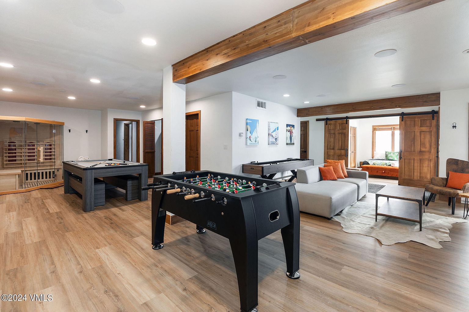 This is a spacious basement featuring a variety of entertainment options. A foosball table is prominently displayed, along with a pool table and a shuffleboard table. The room also includes a seating area with a gray sectional sofa and a cowhide rug, creating a comfortable and inviting space for relaxation and recreation.
