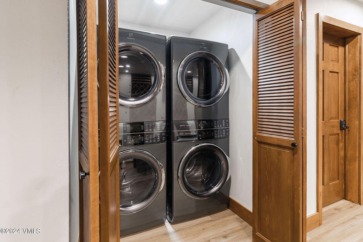 This image showcases a well-organized laundry room featuring stacked Electrolux washer and dryer units in a dark gray finish. The units are neatly tucked into a closet space with louvered wooden doors, adding a touch of rustic charm. The light wood flooring complements the overall clean and functional design of the laundry area.