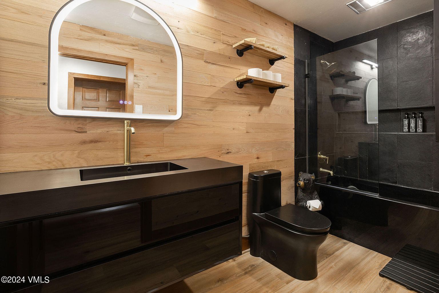 This is a modern bathroom featuring a black vanity with a dark sink and gold faucet, complemented by a wood-paneled wall and an arched mirror with integrated lighting. A black toilet and a decorative bear statue add character, while the shower area showcases dark tiles and glass enclosure. The overall design is sleek and contemporary with a rustic touch.