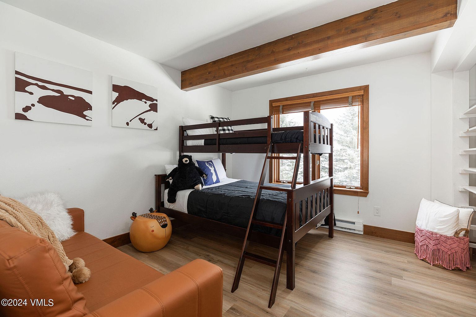 This is a cozy guest bedroom featuring a wooden bunk bed with black bedding and decorative pillows. The room is decorated with modern art and a playful bear plush toy, adding a touch of whimsy. Natural light streams in through a window, complementing the light wood flooring and creating a warm, inviting atmosphere.
