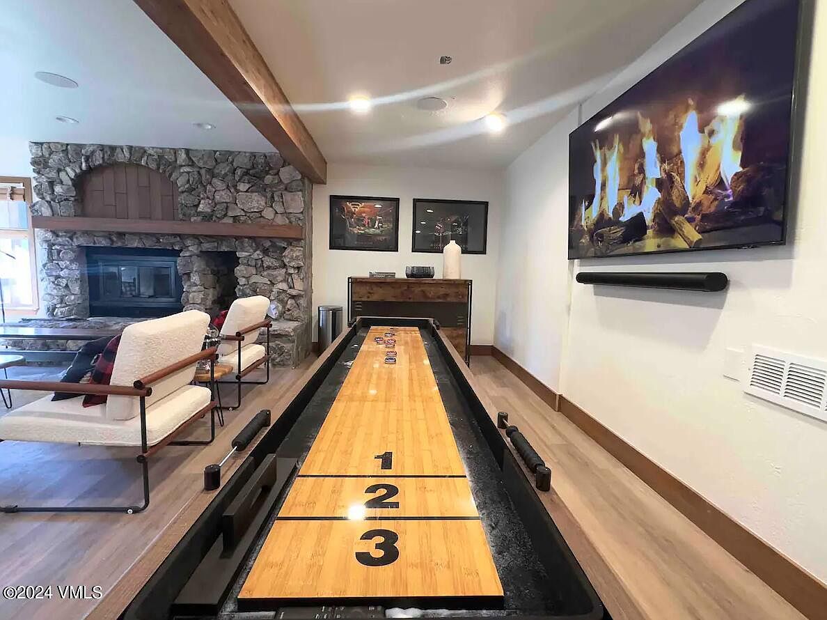 This interior shot showcases a living room featuring a stone fireplace, comfortable seating, and a shuffleboard table. A large television is mounted on the wall, displaying a fireplace scene, adding to the cozy ambiance. The room is well-lit and appears to be designed for relaxation and entertainment.