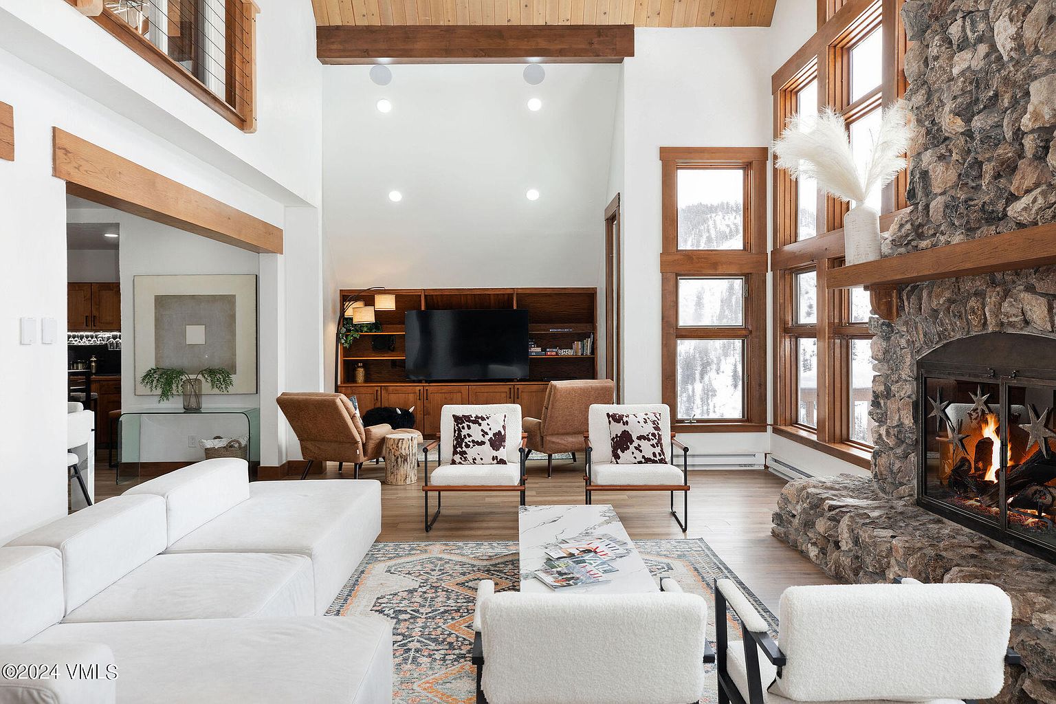 This is a spacious living room featuring a stone fireplace, large windows with wooden frames, and a high ceiling with wooden beams. The room is furnished with a white sectional sofa, accent chairs, and a patterned area rug, creating a cozy and inviting atmosphere. The overall style is modern rustic with a focus on natural materials and textures.