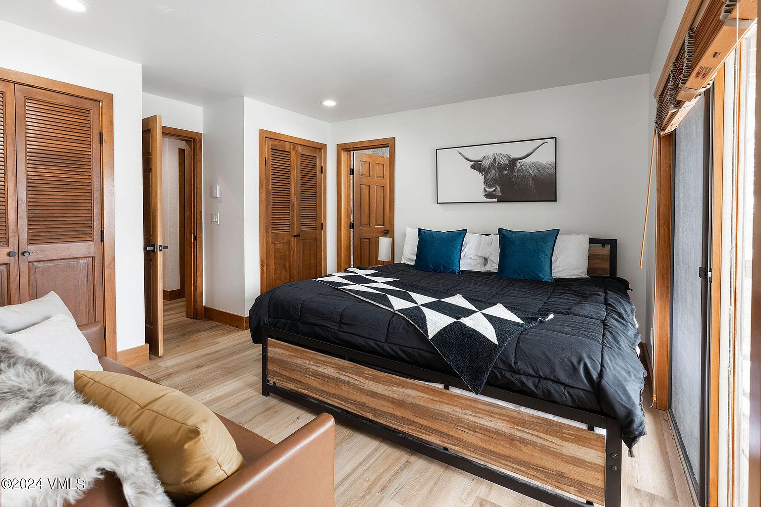 This is a bedroom featuring a wooden-framed bed with a black comforter, adorned with a black and white patterned throw. Two blue pillows and two white pillows are placed against the headboard. A black and white photograph of a bull hangs above the bed. The room has wooden floors and doors, and a sliding glass door provides natural light.