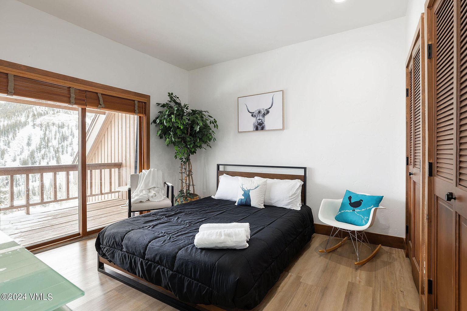 This is a cozy primary bedroom featuring a black bedspread, white pillows, and a decorative pillow with a deer design. A large window provides natural light and a view of a snowy landscape. The room is decorated with a potted tree and a framed picture of a highland cow, creating a serene and inviting atmosphere.
