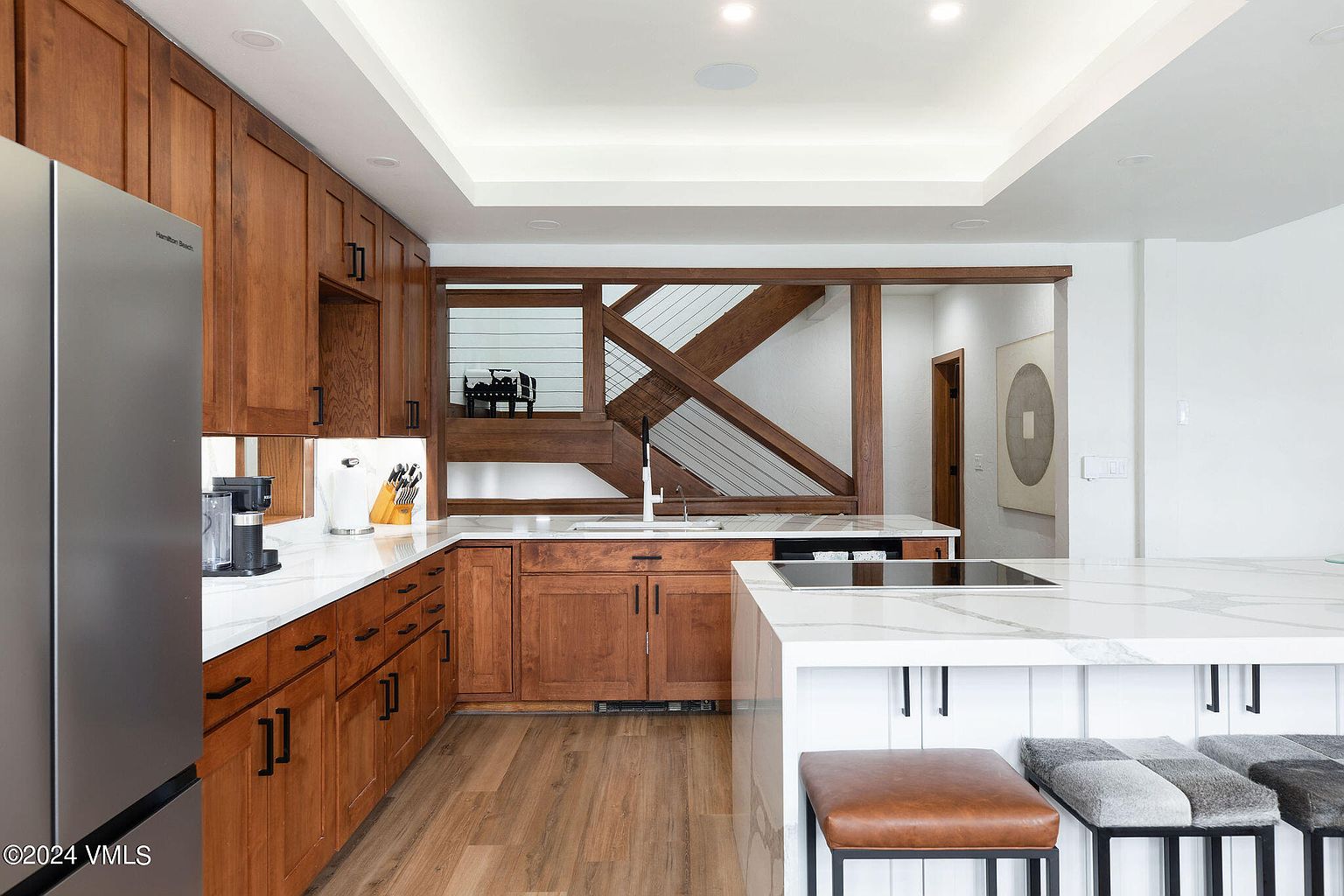 This is a well-lit kitchen featuring wooden cabinetry with black hardware and a white countertop. A stainless steel refrigerator is visible on the left, and a kitchen island with seating is in the foreground. The kitchen has a modern design with a unique wooden partition in the background.