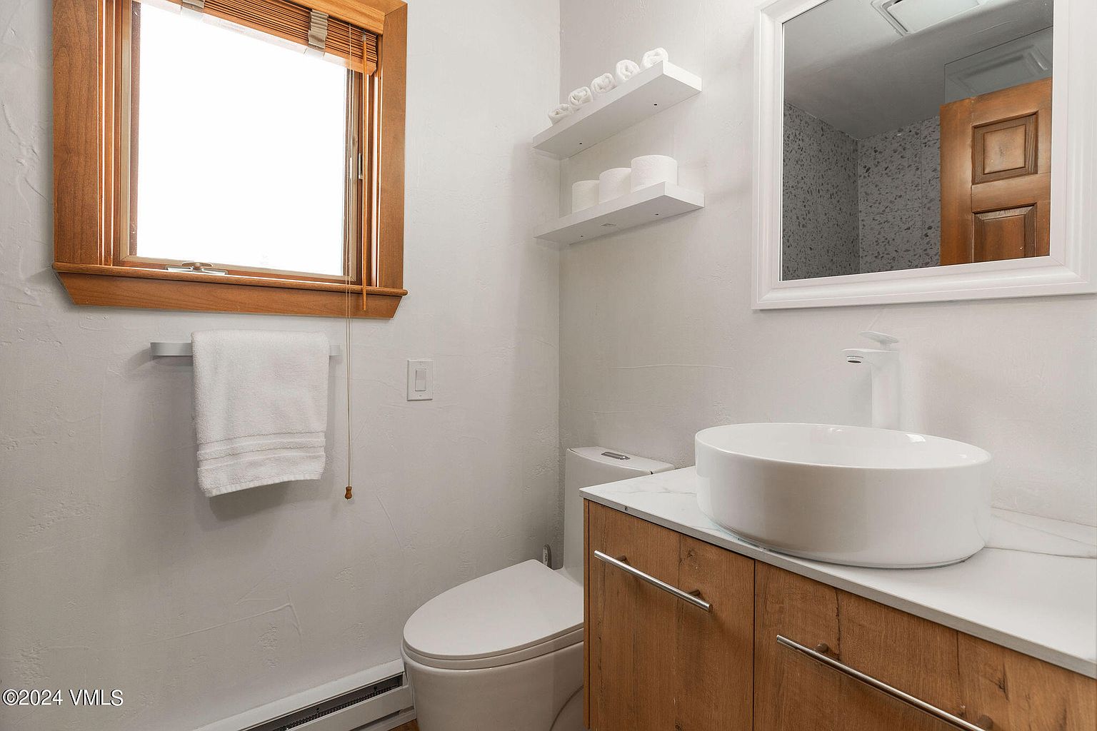 This is a well-lit guest bathroom featuring a modern vessel sink atop a wooden vanity with sleek, silver hardware. A white toilet sits adjacent to the vanity, and floating shelves above it hold neatly arranged towels and toilet paper. The room is painted in a light color, enhancing the clean and minimalist aesthetic.