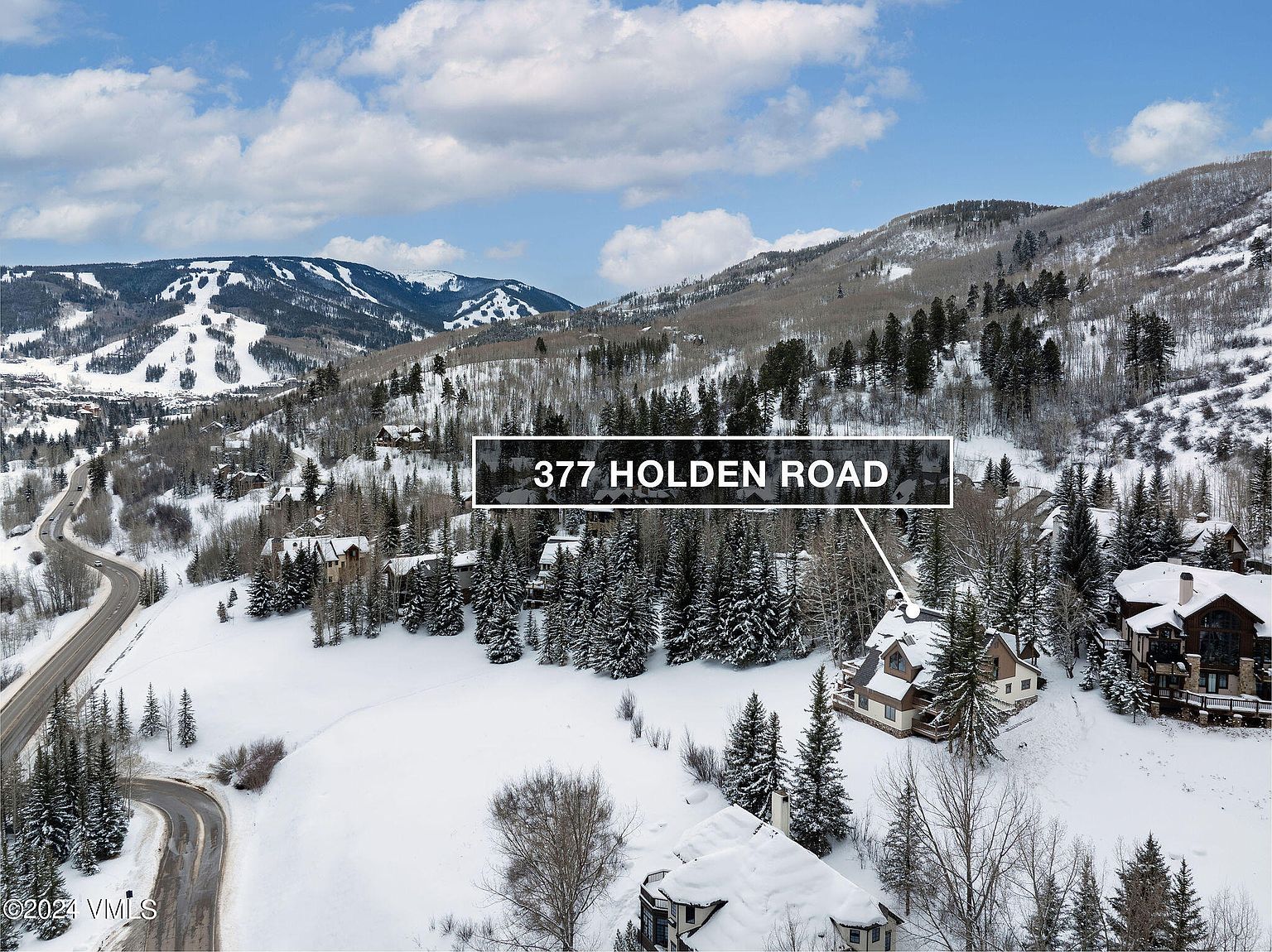 This aerial view showcases a beautiful property nestled in a snowy, mountainous landscape. The house, with its unique architecture, is surrounded by evergreen trees and a blanket of snow, creating a serene and picturesque setting. A winding road leads to the property, suggesting privacy and seclusion.
