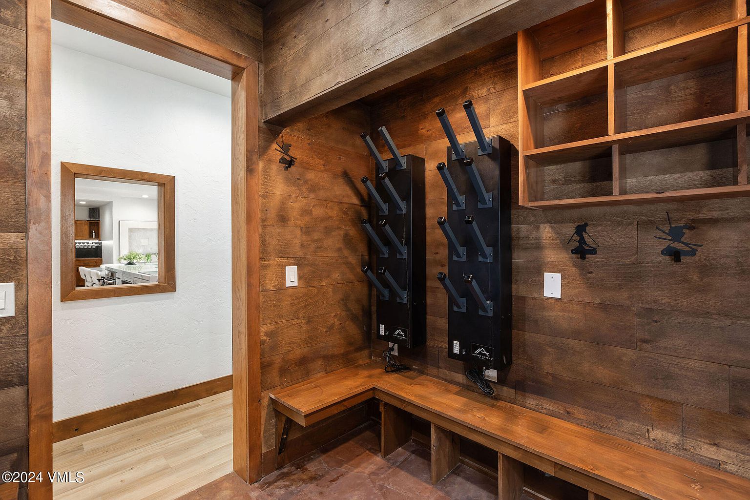 This interior shot showcases a rustic-style hallway or entryway with wooden walls and trim. A built-in bench with storage underneath sits beneath ski boot dryers, and open shelving adds to the functional design. A doorway leads to another room, visible through a framed mirror, creating a sense of depth and connection within the home.