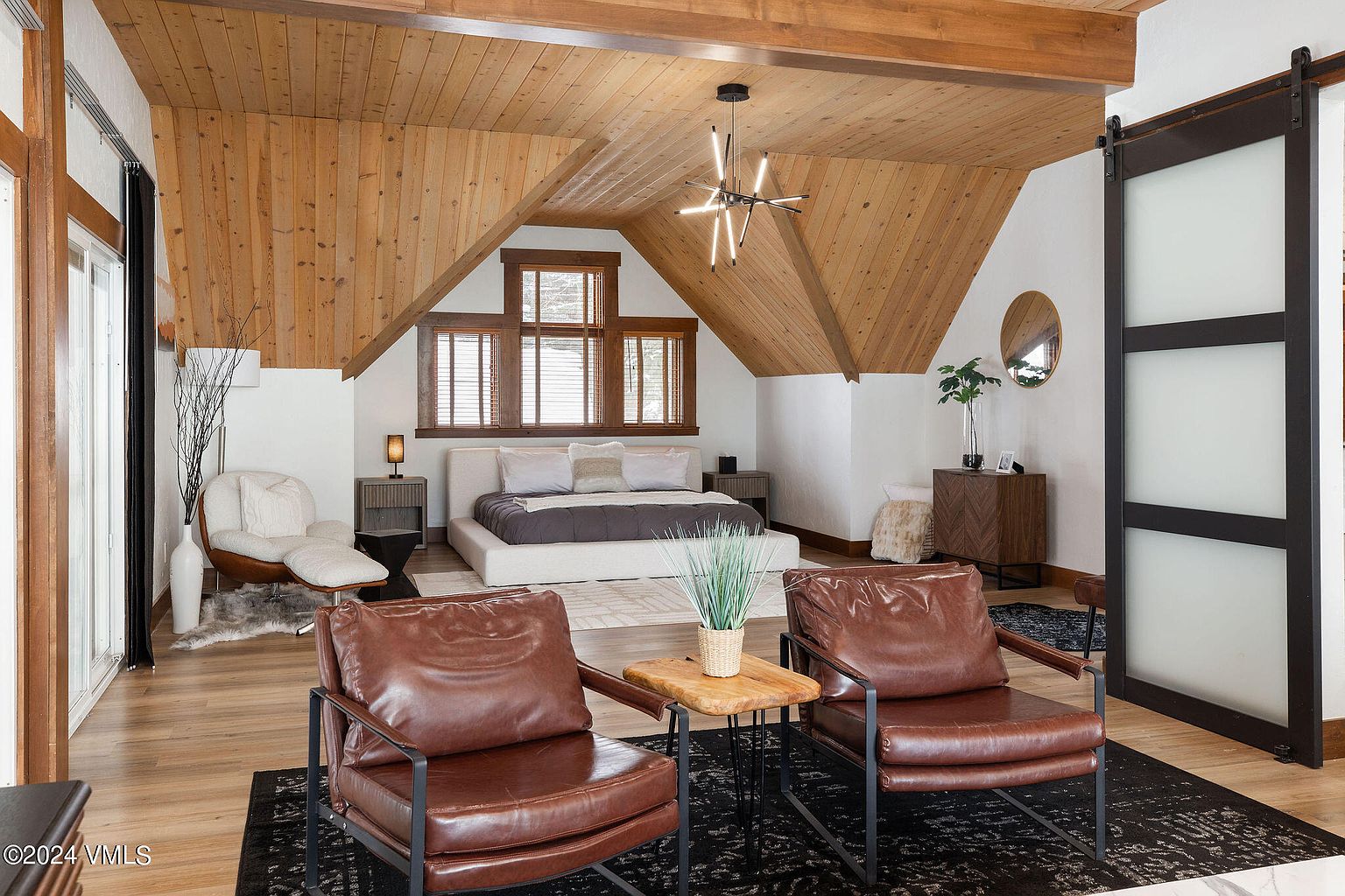 This is a cozy primary bedroom featuring a wood-paneled ceiling and an accent wall. The room is furnished with a platform bed, two leather chairs, and a modern light fixture. The overall style is rustic and modern, creating a warm and inviting atmosphere.