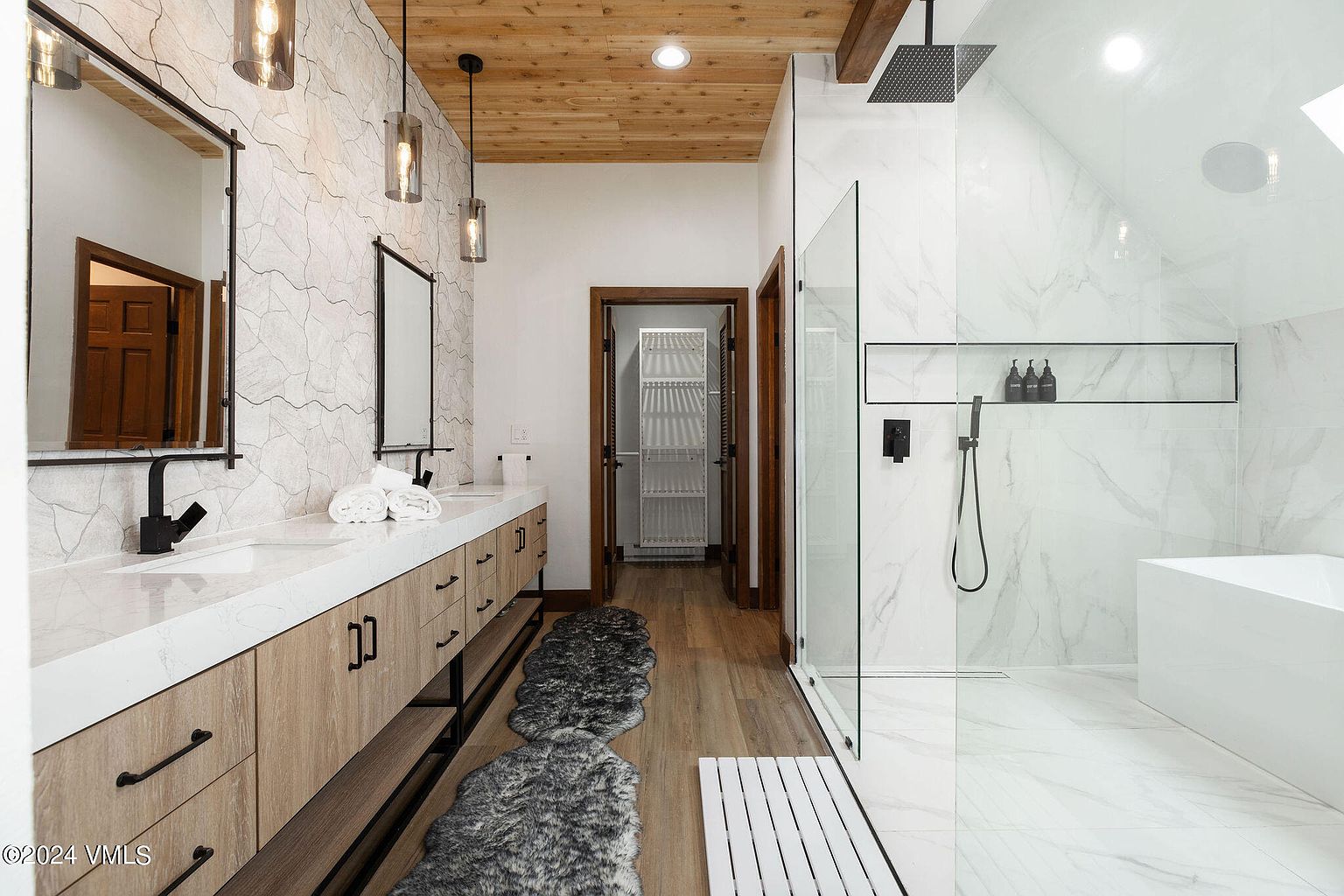 This is a primary bathroom featuring a double vanity with a white countertop and light wood cabinets, complemented by black hardware and modern pendant lighting. A spacious walk-in shower with glass enclosure and a separate soaking tub are visible, all finished with marble-like tiling. The overall design is modern and luxurious, creating a spa-like atmosphere.