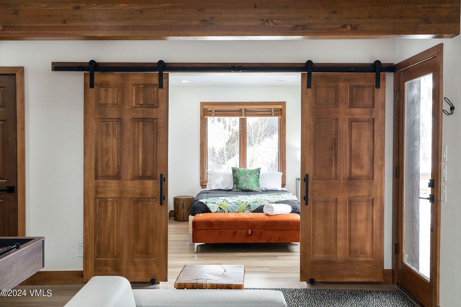 This is an interior shot of a primary bedroom, viewed through a set of rustic sliding barn doors. The bedroom features a bed with white linens and a patterned throw, a window with wooden blinds, and an orange ottoman at the foot of the bed. The style is a blend of rustic and modern, creating a cozy and inviting atmosphere.