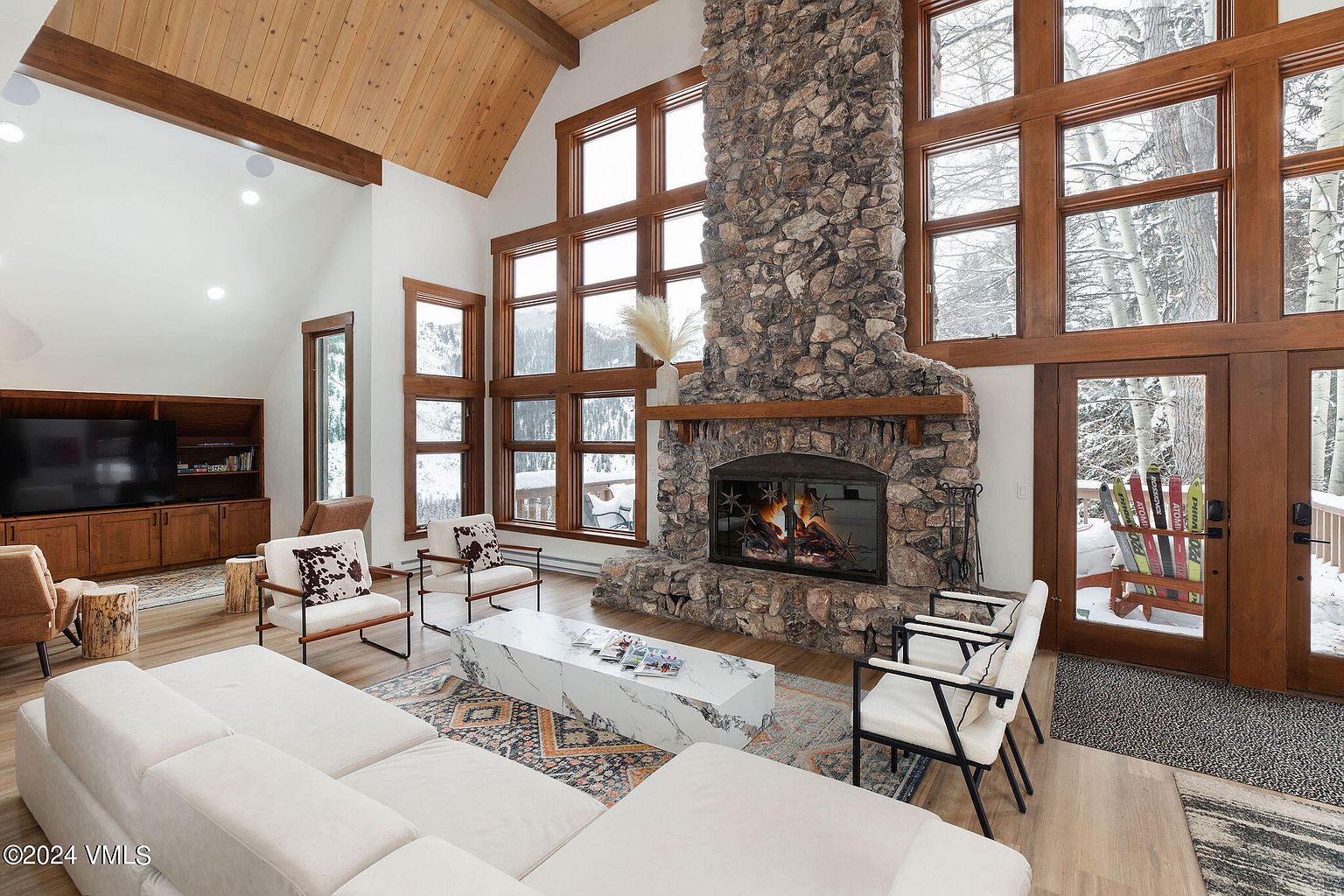 This is an interior shot of a luxurious living room featuring a grand stone fireplace as a focal point, complemented by large windows offering snowy outdoor views. The room is furnished with a comfortable white sectional sofa, modern chairs, and a stylish marble coffee table, all set on a patterned rug. The high, wood-paneled ceiling adds to the room's spacious and elegant atmosphere.