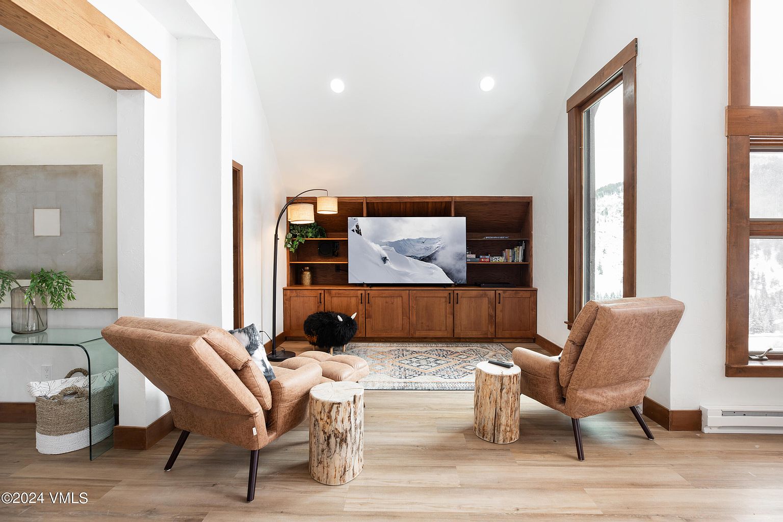 This inviting living room features a warm and cozy atmosphere with two leather armchairs facing a wooden entertainment center. A patterned rug anchors the space, while natural light streams in through the windows, highlighting the wood flooring. The room is tastefully decorated with rustic accents, creating a comfortable and stylish living space.