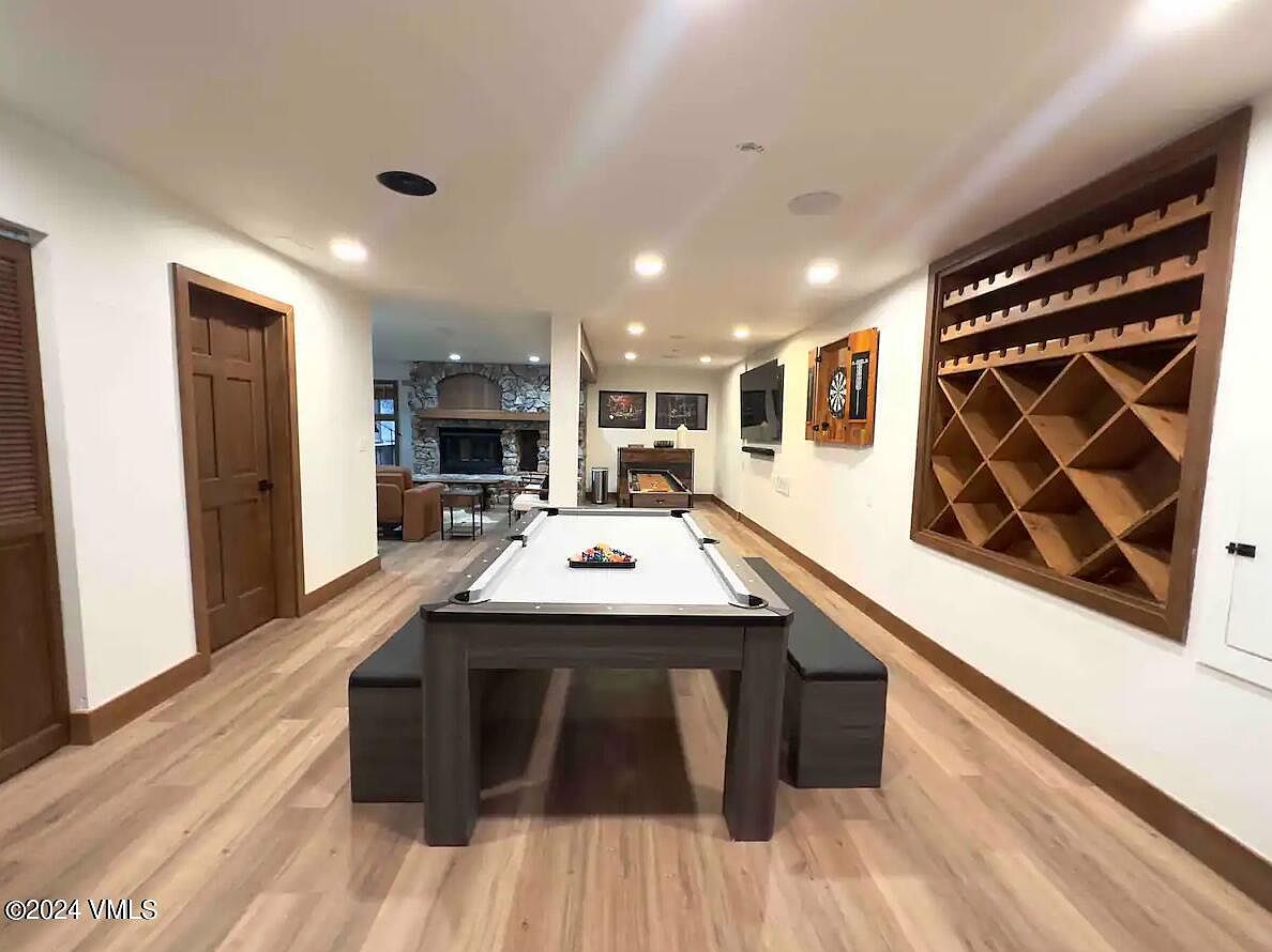 This is a well-lit basement featuring a pool table with benches, a wine rack, and a dartboard. The room has light wood flooring and white walls with wood trim, creating a warm and inviting atmosphere. An open doorway leads to another living space with a fireplace, suggesting a spacious and versatile area for entertainment and relaxation.