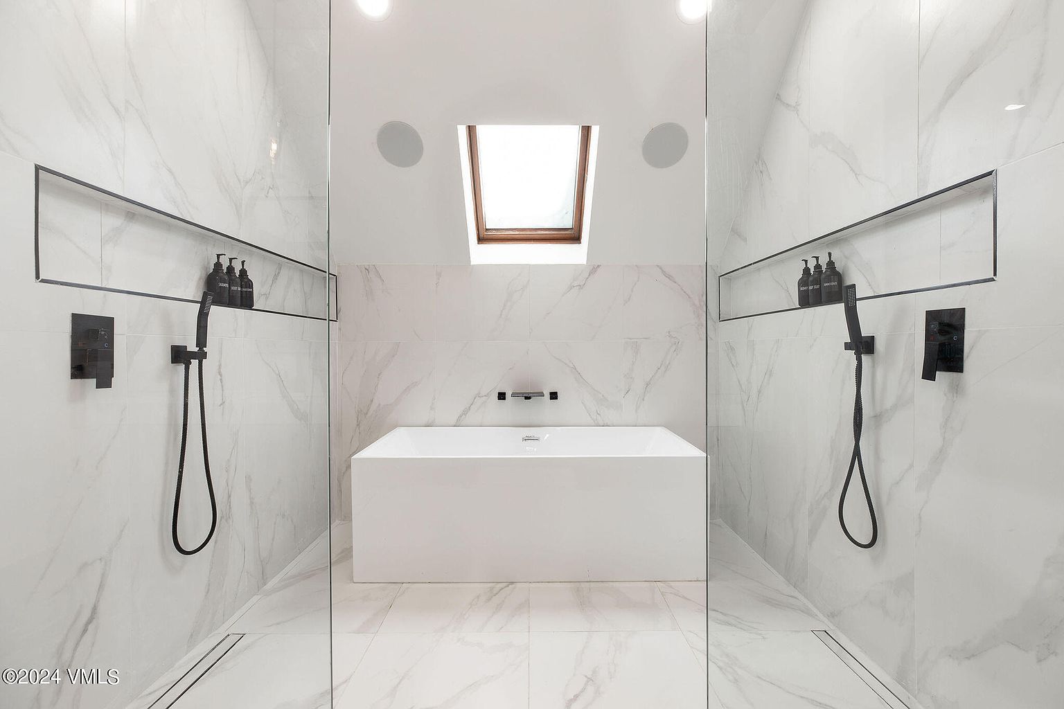 This is a modern primary bathroom featuring white marble walls and floors, creating a luxurious and clean aesthetic. The bathroom includes a freestanding white bathtub centered beneath a skylight, flanked by glass-enclosed shower areas on either side, each equipped with black fixtures and recessed shelving. The design emphasizes a minimalist and spa-like atmosphere.