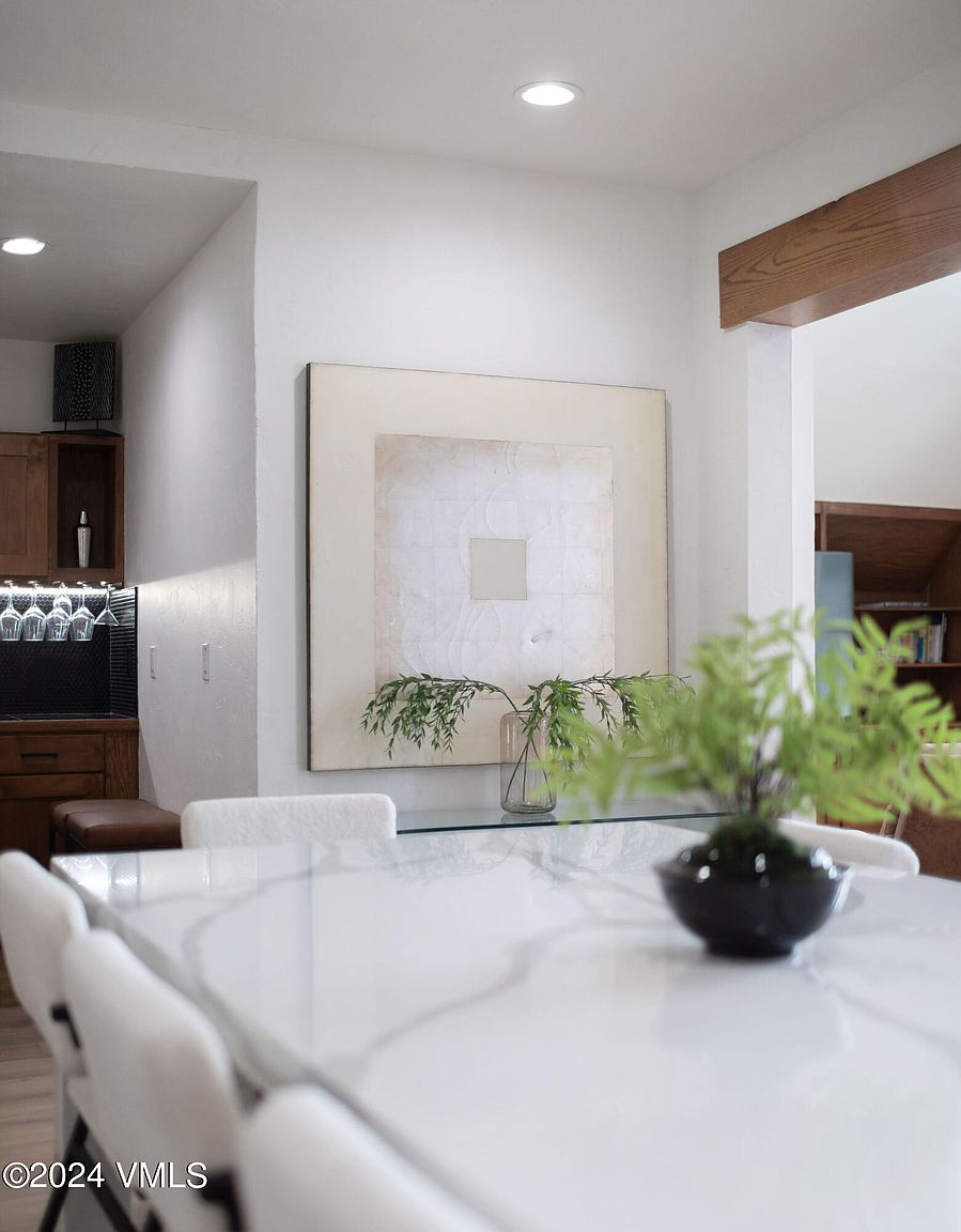 This interior shot showcases a dining room with a modern aesthetic. A large, white marble-topped table dominates the foreground, surrounded by white chairs. Artwork hangs on the wall, complemented by decorative greenery, creating an inviting and stylish dining space.