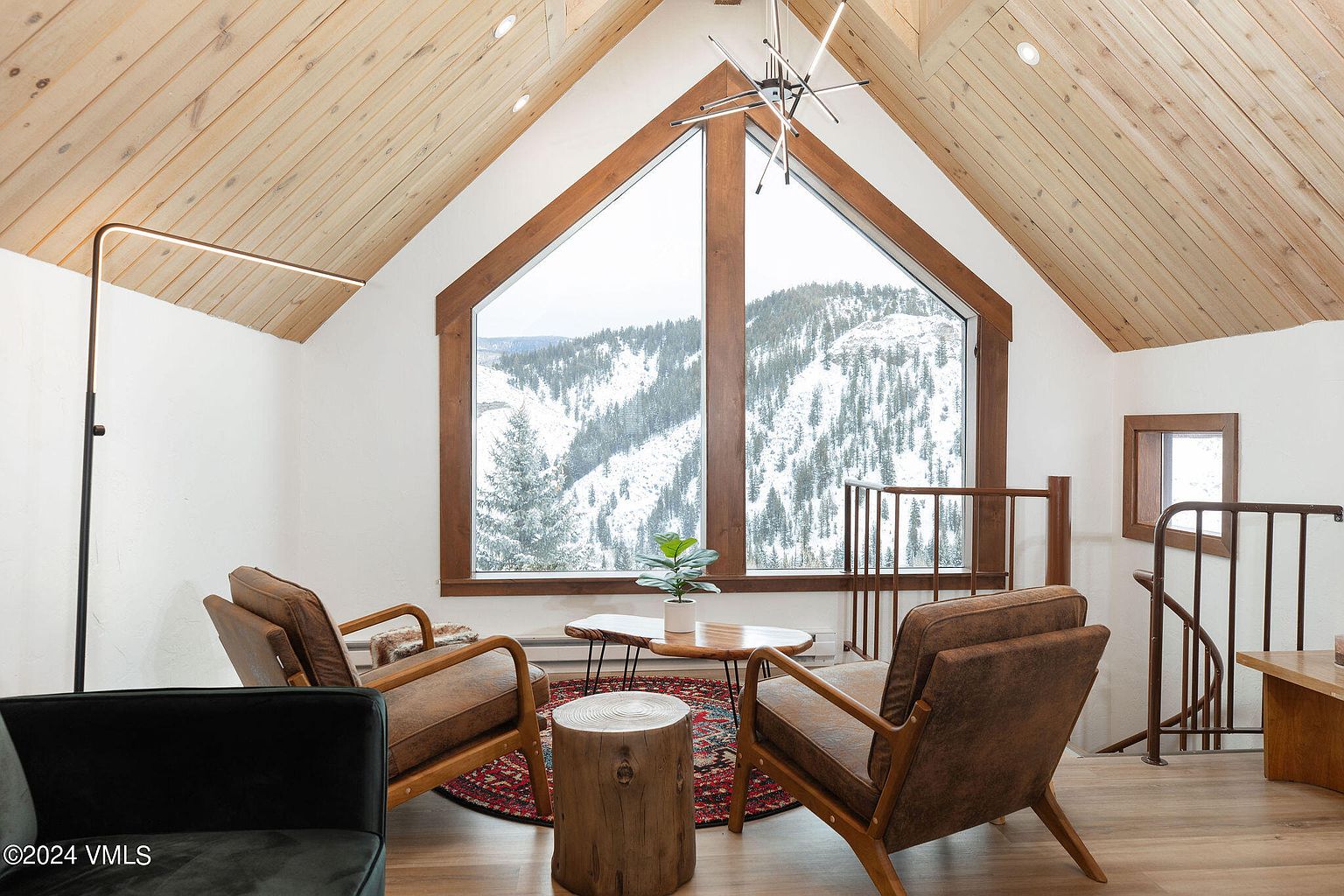 This inviting living room features a striking A-frame window showcasing a snow-covered mountain view, complemented by warm wood accents on the ceiling and window frame. Two mid-century modern chairs flank a rustic wooden coffee table and stool, set upon a patterned rug, creating a cozy and stylish space. A modern floor lamp and chandelier add to the room's contemporary charm.