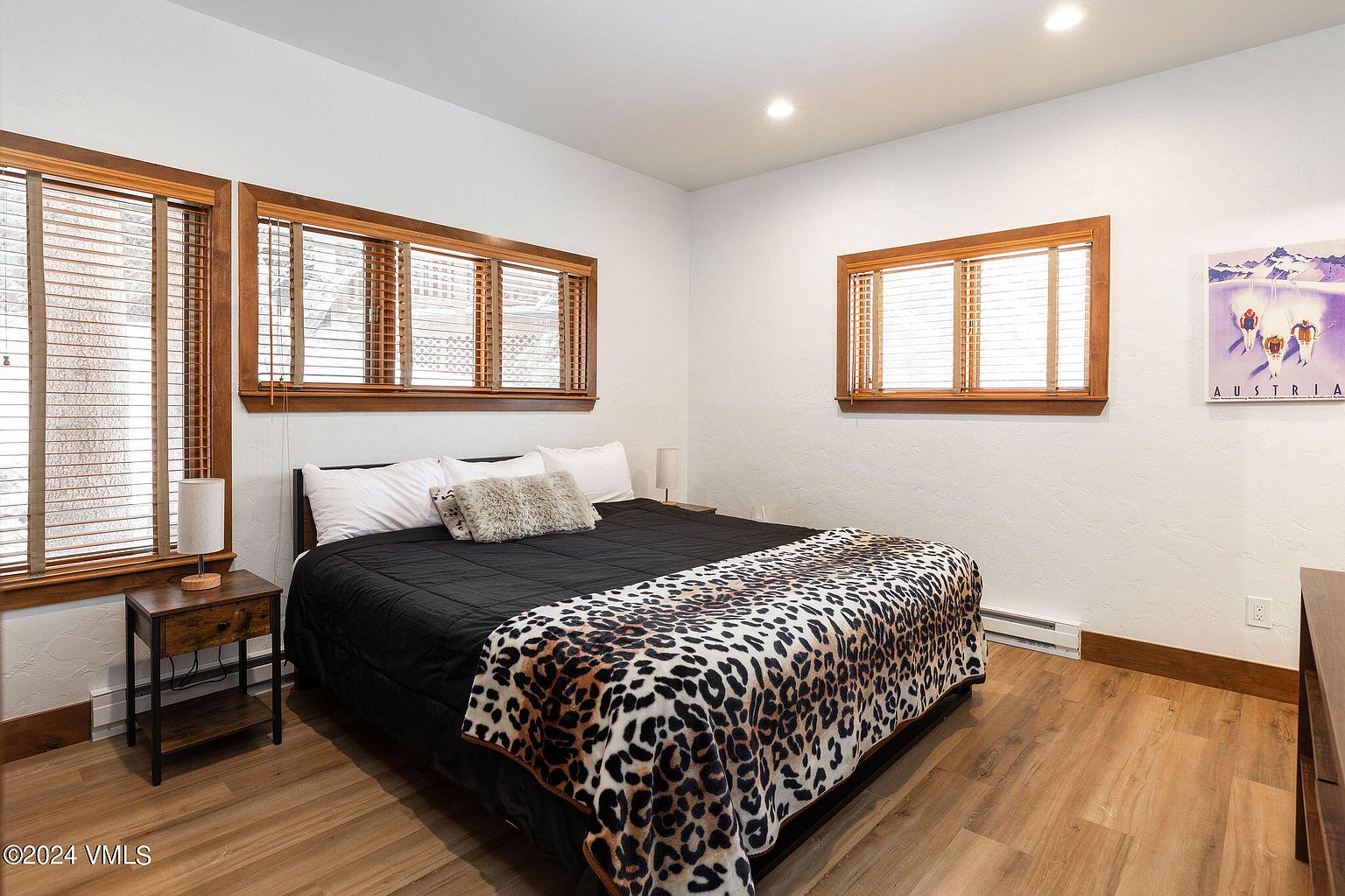 This is a cozy bedroom featuring a bed with a black comforter and a leopard print throw blanket. The room has three windows with wooden frames and blinds, allowing natural light to filter in. A wooden nightstand with a lamp sits beside the bed, and the flooring is light-colored wood, creating a warm and inviting atmosphere.