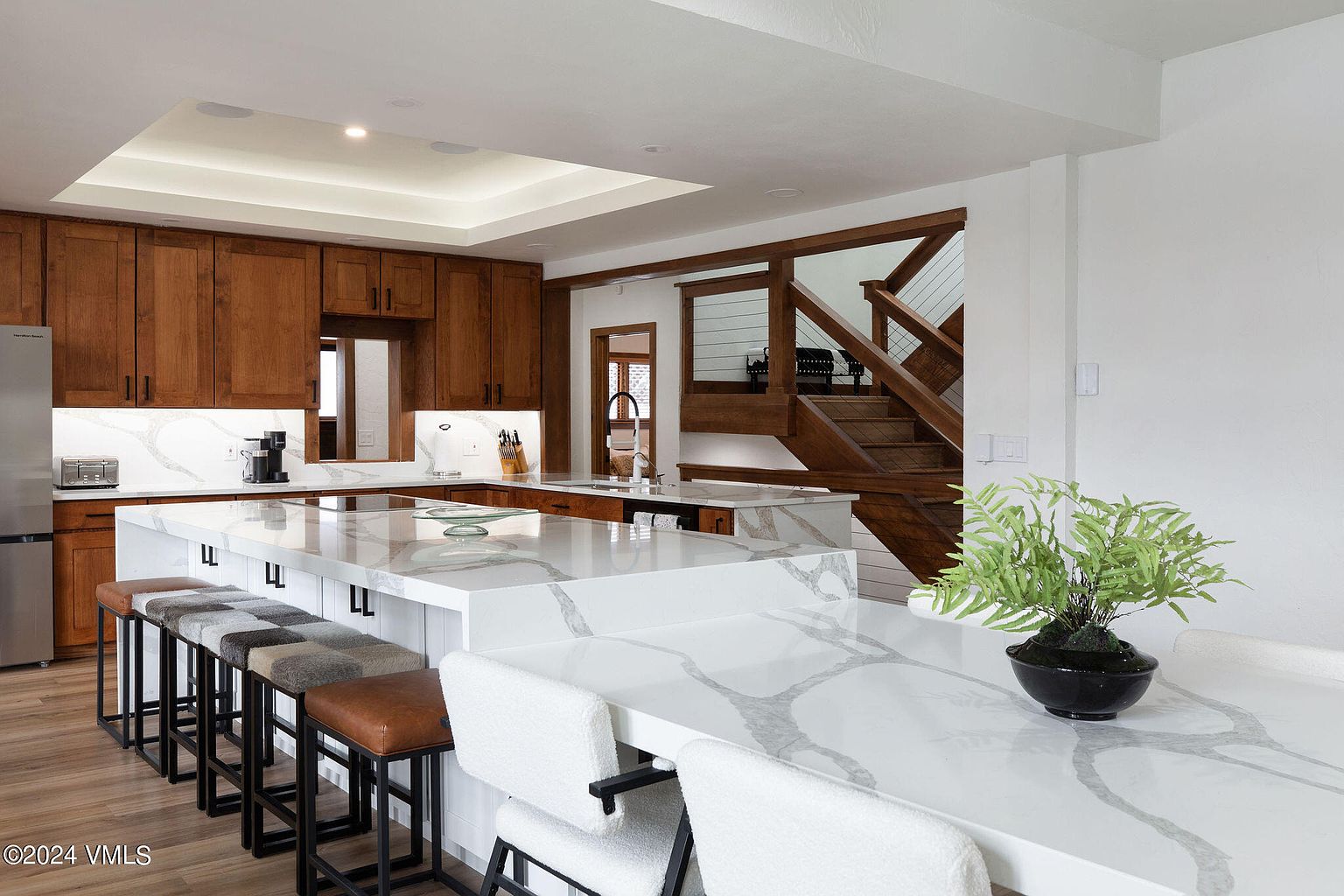 This is a well-lit kitchen featuring wooden cabinets, stainless steel appliances, and a large island with a white countertop with gray veining. The kitchen also has a staircase leading to another floor, adding architectural interest. The overall impression is modern and inviting.