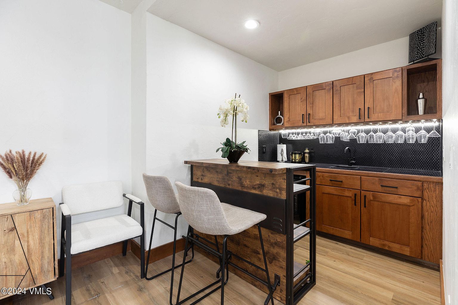 This interior shot showcases a stylish bar area within a kitchen setting, featuring wooden cabinetry, a black tiled backsplash, and a rustic-modern bar counter with two stools. The space is well-lit, highlighting the warm tones of the wood and the sleek black accents, creating an inviting and sophisticated atmosphere. A white chair and wooden cabinet are on the left side of the image.