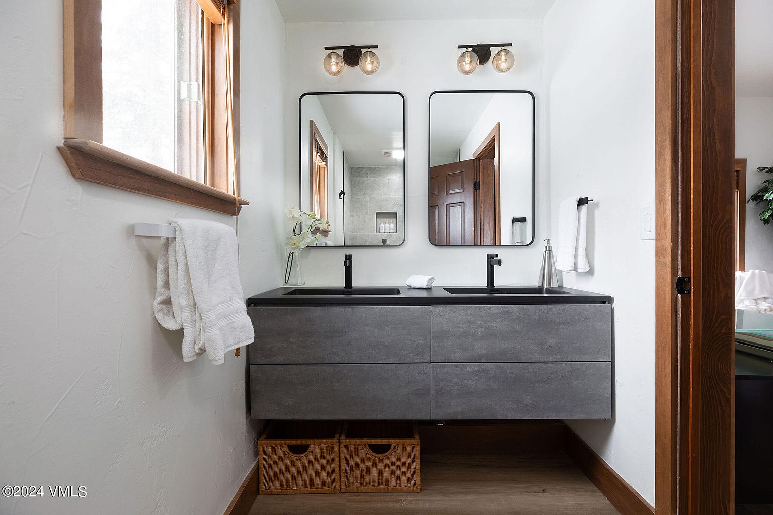 This is a well-lit bathroom featuring a modern double vanity with a gray textured finish and black countertops. Two rectangular mirrors hang above the sinks, each illuminated by a stylish light fixture. Wicker baskets provide storage underneath the vanity, and the overall design is clean and contemporary.