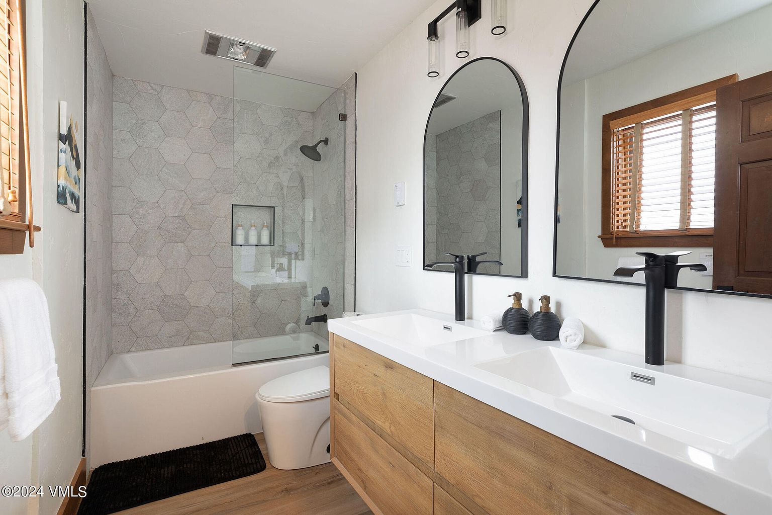 This is a well-lit bathroom featuring a double sink vanity with a light wood finish and white countertop. Above the vanity are two arched mirrors with black frames, complemented by modern black faucets and soap dispensers. The shower/tub area is enclosed with glass and features hexagonal tiling, adding a contemporary touch to the space.
