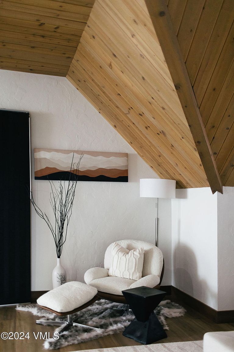 This interior shot showcases a cozy living room nook with a modern aesthetic. The space features a comfortable armchair with a matching ottoman, complemented by a black geometric side table and a stylish area rug. A piece of artwork hangs above the chair, and the room is illuminated by a floor lamp, creating a warm and inviting atmosphere.