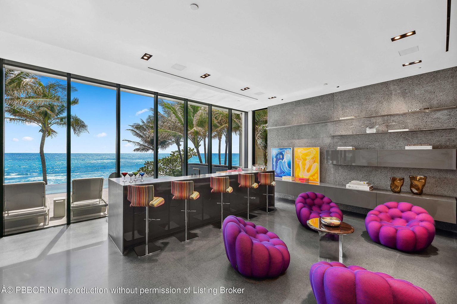 This interior shot showcases a modern living room with a stunning ocean view through large windows. The room features a sleek bar area with stools, unique purple seating, and a gray accent wall with built-in shelving. The polished floors and contemporary lighting contribute to the room's luxurious and stylish ambiance.