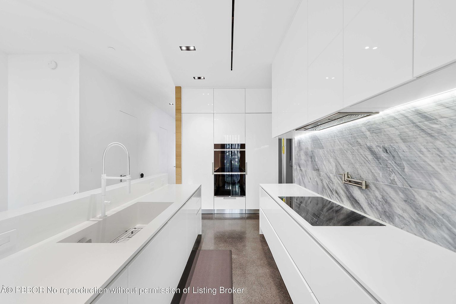 This is a modern, minimalist kitchen featuring sleek white cabinetry and countertops. The kitchen is well-lit with recessed lighting and features high-end appliances, including built-in ovens and a cooktop. The backsplash is a striking marble, adding a touch of luxury to the clean design.