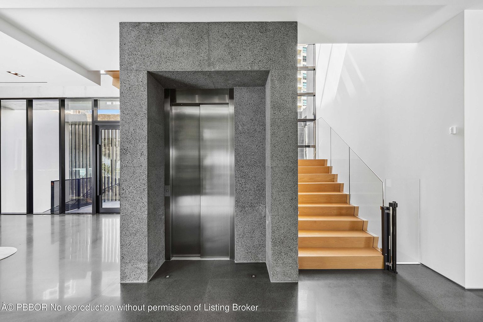This interior shot showcases a modern hallway with a unique architectural design. A sleek, stainless steel elevator is framed by textured gray walls, contrasting with the warm wood stairs featuring glass railings. The polished concrete floors reflect light, enhancing the contemporary aesthetic of the space.