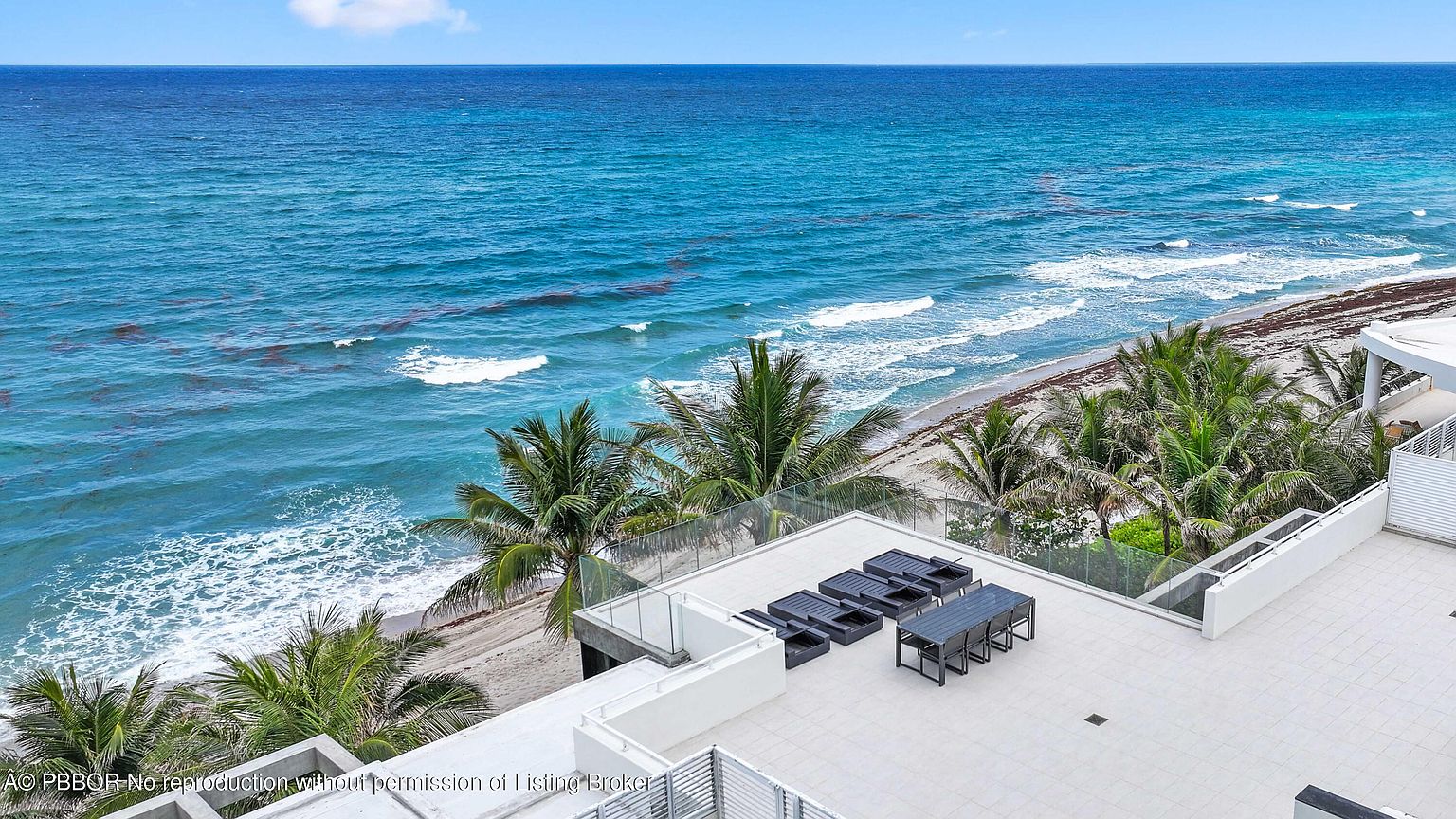 This image showcases a luxurious rooftop patio/deck/balcony with modern outdoor furniture, including lounge chairs and a dining set. The space is surrounded by glass railings, offering unobstructed panoramic views of the ocean and beach. Palm trees add a tropical touch, enhancing the overall upscale and serene atmosphere.