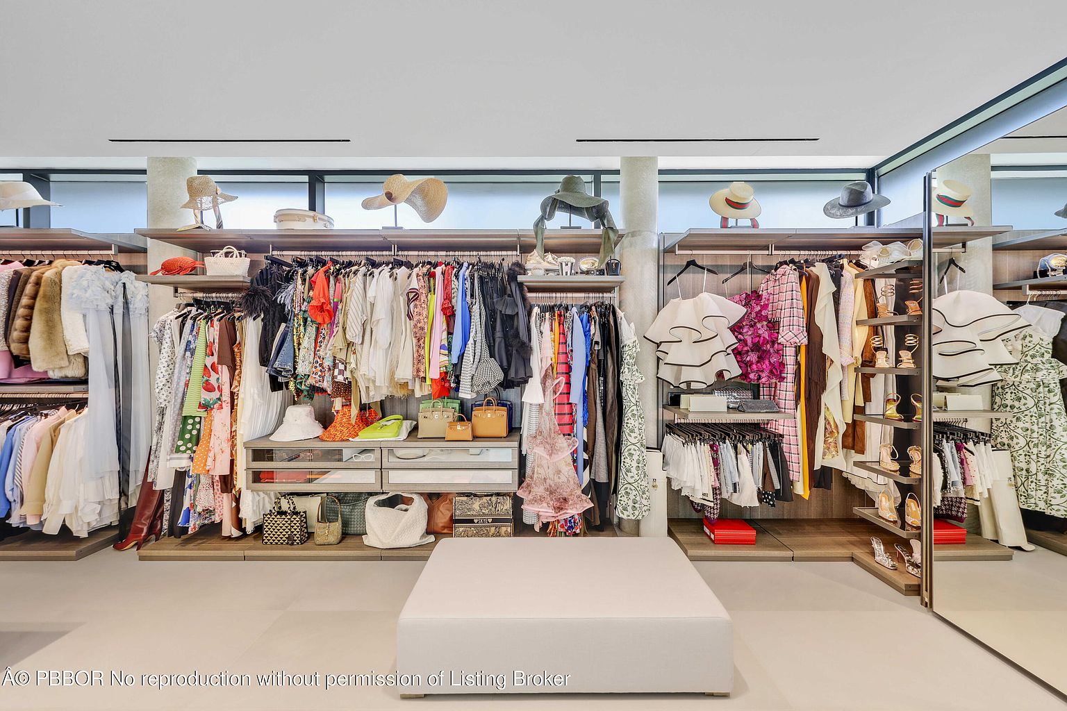 This is a well-organized walk-in closet featuring an extensive collection of clothing, shoes, and accessories. The closet is designed with multiple shelves, hanging rods, and drawers, providing ample storage space. A central ottoman offers a seating area, and the overall design is luxurious and functional, appealing to those seeking a high-end storage solution.