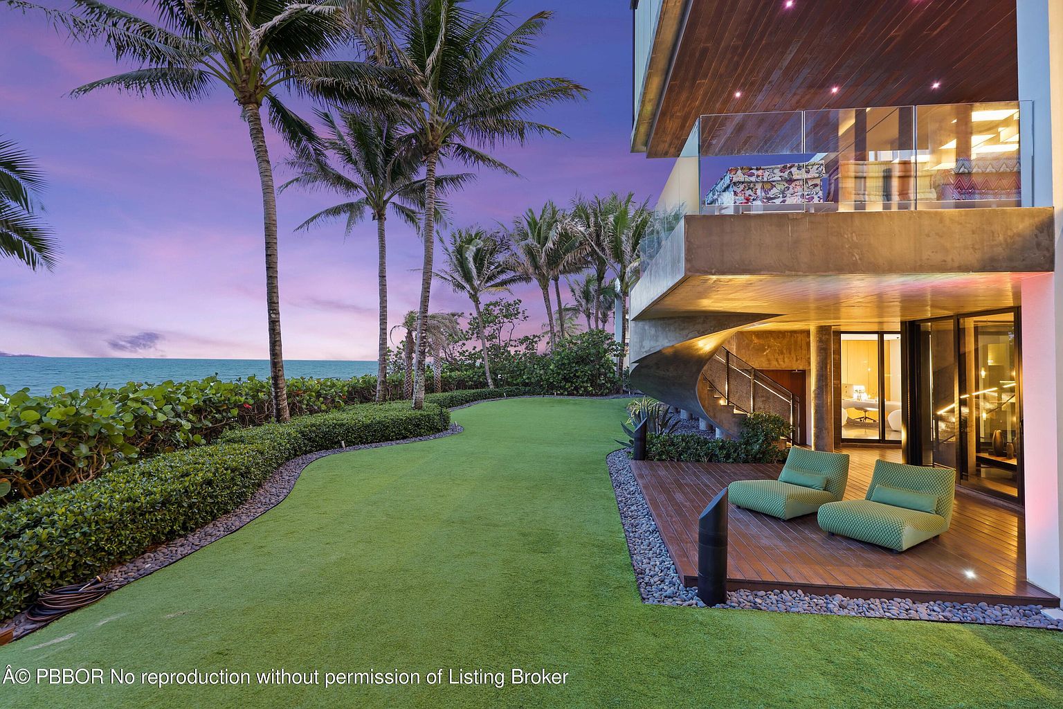 This exterior rear view showcases a modern home with a lush green lawn extending to the ocean. Palm trees line the property, and a unique spiral staircase leads to a deck with comfortable seating. The architecture is contemporary, blending seamlessly with the natural landscape.