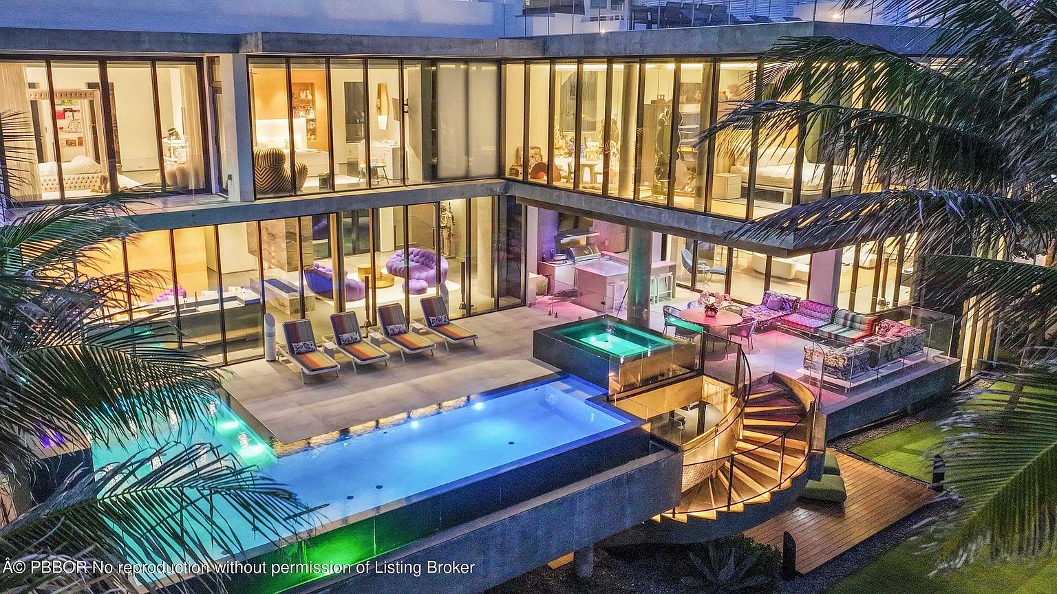 This image showcases a luxurious modern home's exterior, emphasizing its stunning pool and spa area. The pool features a unique design with illuminated water, complemented by a nearby hot tub. A spiral staircase leads to an upper-level patio with stylish outdoor furniture, all set against a backdrop of lush tropical foliage and a contemporary architectural design.