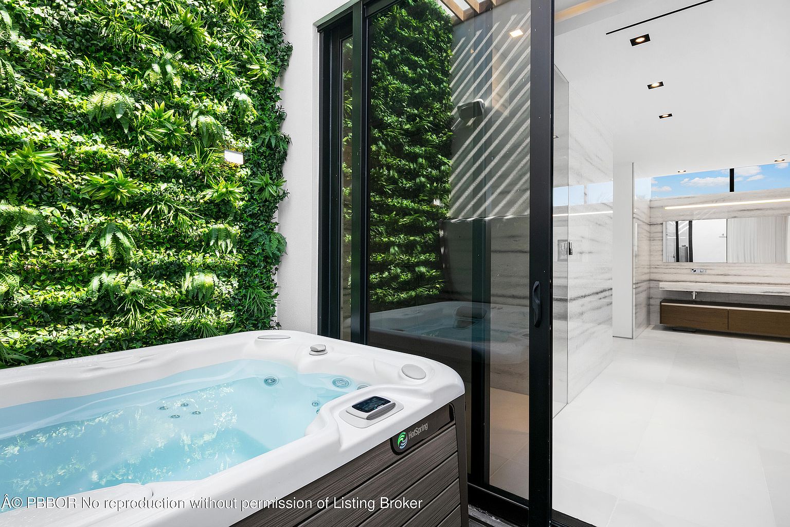 This image showcases a luxurious outdoor spa area featuring a modern hot tub surrounded by a vibrant green wall. Sliding glass doors provide seamless access to an elegant bathroom with marble accents and contemporary fixtures. The scene exudes relaxation and sophistication, highlighting the property's high-end amenities.