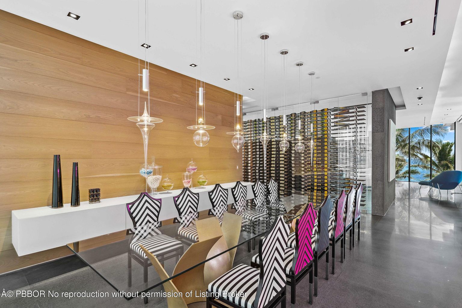 This is a modern dining room featuring a glass-top table surrounded by uniquely patterned chairs. The room is illuminated by pendant lights and features a wine cellar wall, adding a touch of sophistication. Large windows offer a view of the ocean, enhancing the luxurious ambiance.