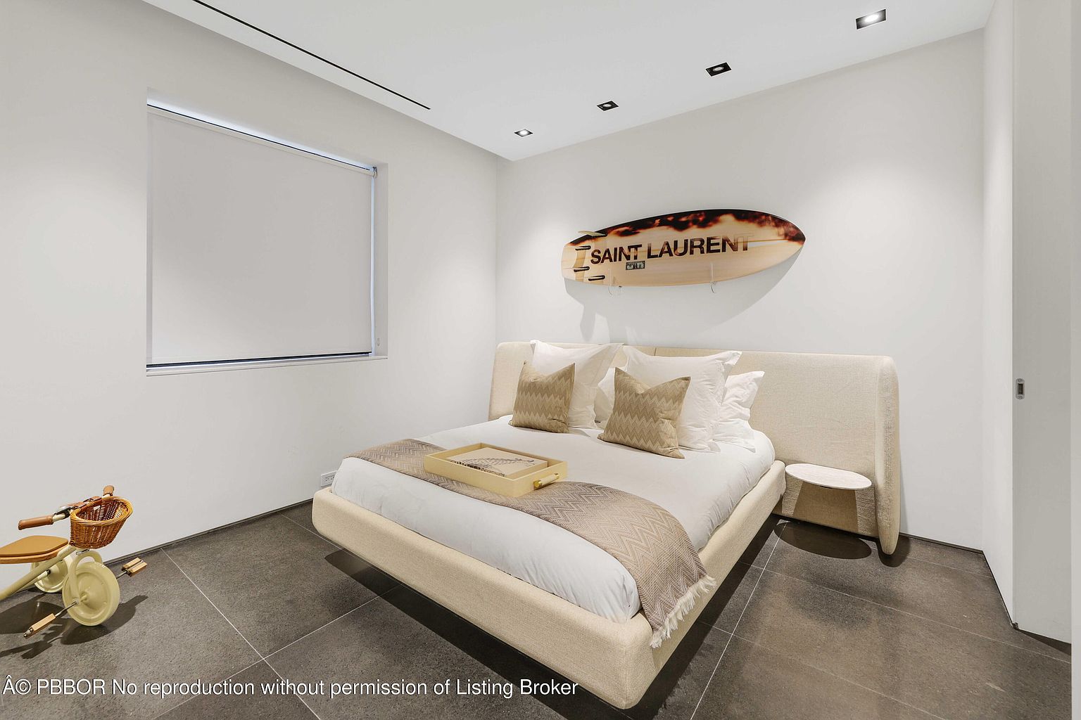 This is a modern bedroom featuring a low-profile platform bed with a light beige upholstered frame and headboard. The room is decorated with a surfboard art piece above the bed, adding a touch of coastal style. The flooring is dark tile, and the walls are painted white, creating a clean and minimalist aesthetic. A child's tricycle is visible in the corner, suggesting the room may be used as a child's bedroom.