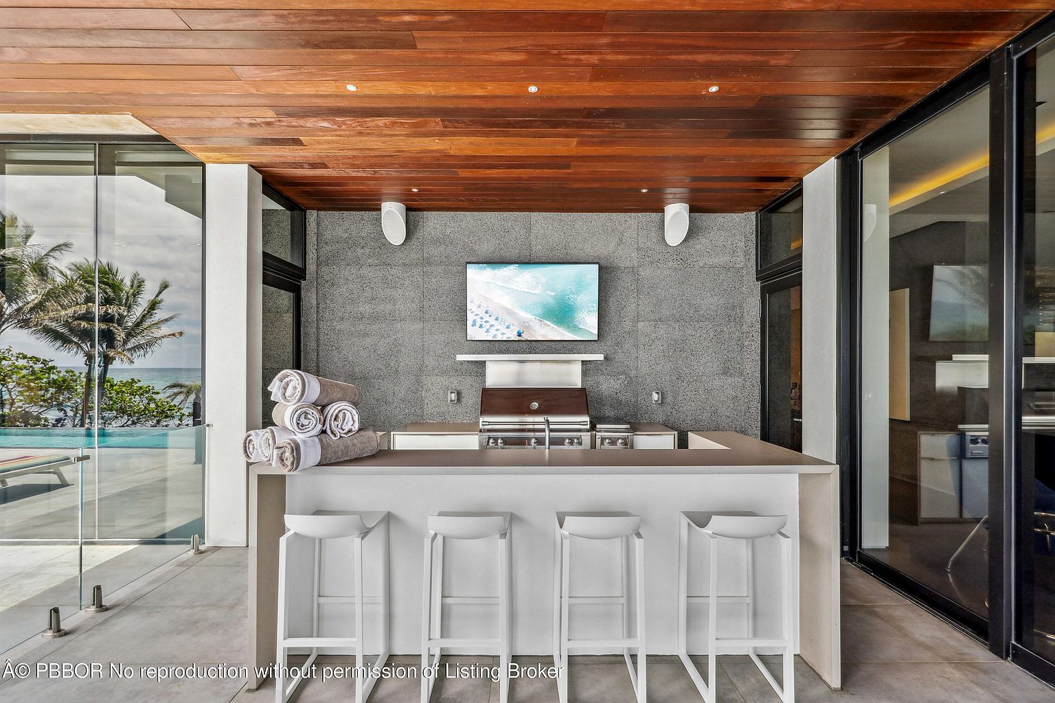 This image showcases an outdoor patio area featuring a built-in grill and bar setup, complete with white bar stools. A flat-screen television is mounted on the wall above the grill, and rolled towels are neatly stacked on the bar counter. The space is partially covered by a wooden ceiling and offers views of the ocean and palm trees, creating a luxurious outdoor living experience.