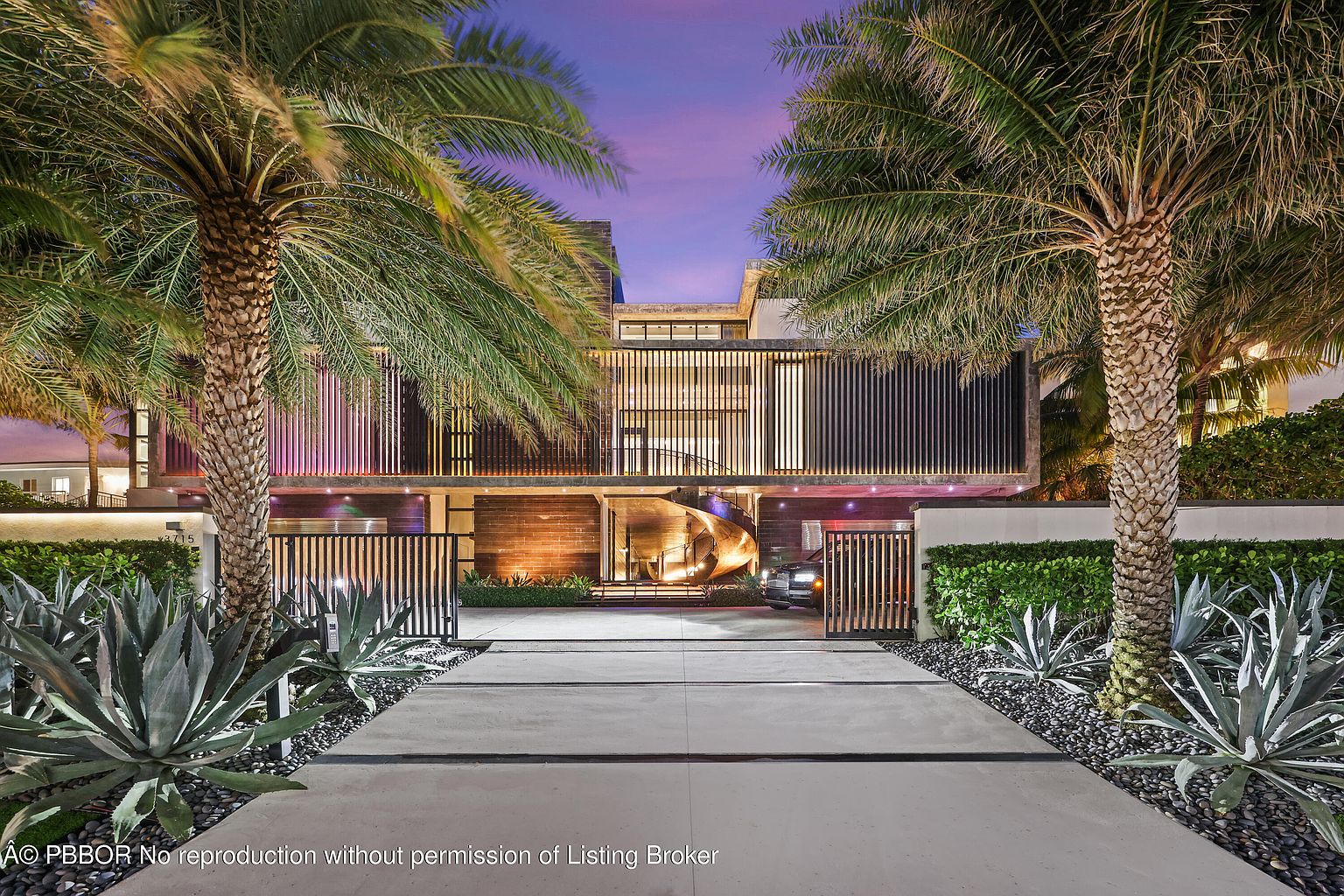 This is a striking front view of a modern luxury home at dusk, framed by two large palm trees. The house features a unique architectural design with horizontal slats and a spiral staircase leading to the entrance. A well-manicured garden with agave plants and a paved driveway add to the property's curb appeal, while a black car is parked in the driveway.