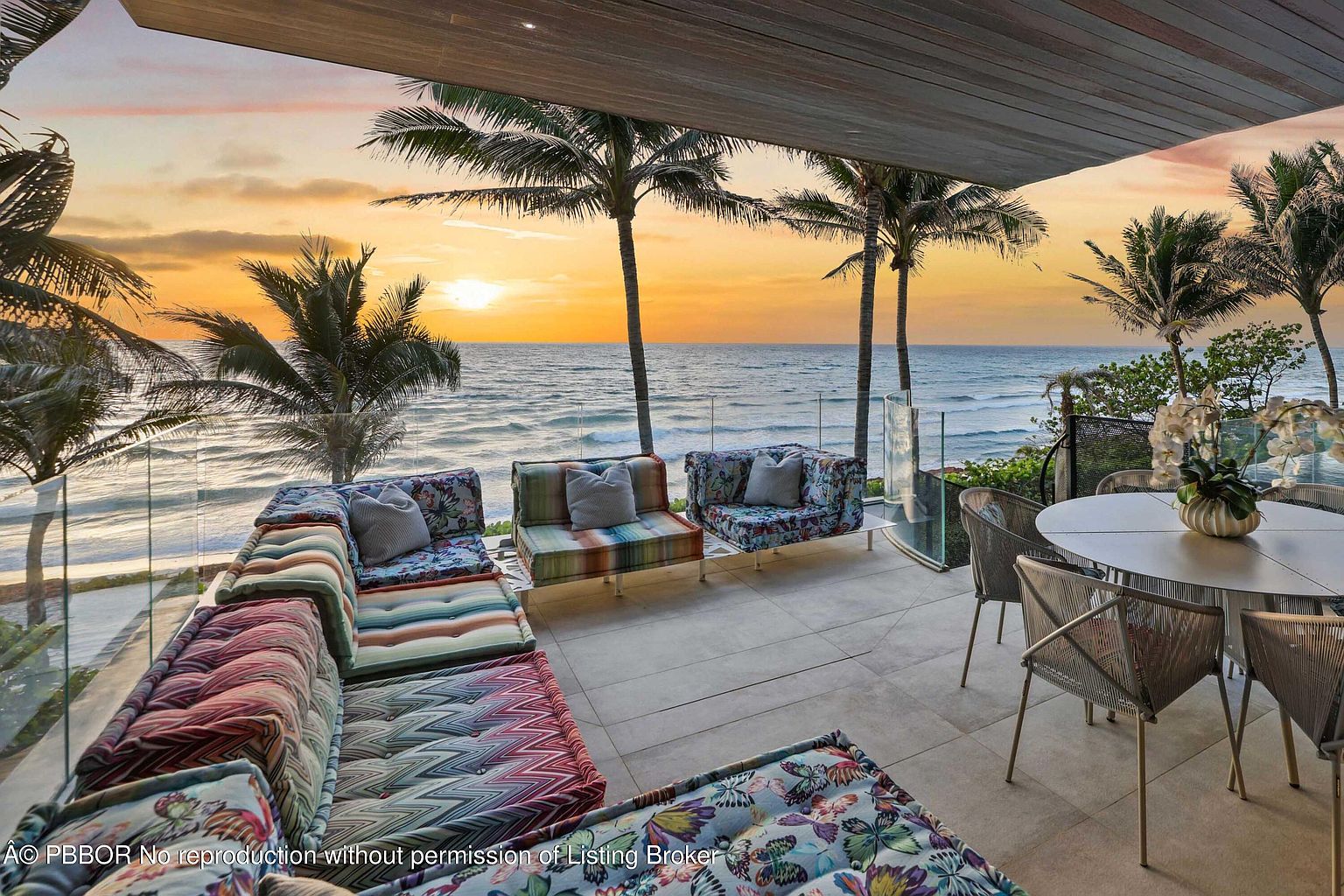 This image showcases a luxurious outdoor patio or balcony with stunning ocean views at sunset. The space is furnished with comfortable, colorful seating arrangements, including a large sectional and individual chairs, complemented by a round dining table with woven chairs. Palm trees frame the picturesque scene, enhancing the tropical ambiance and creating an inviting space for relaxation and entertainment.